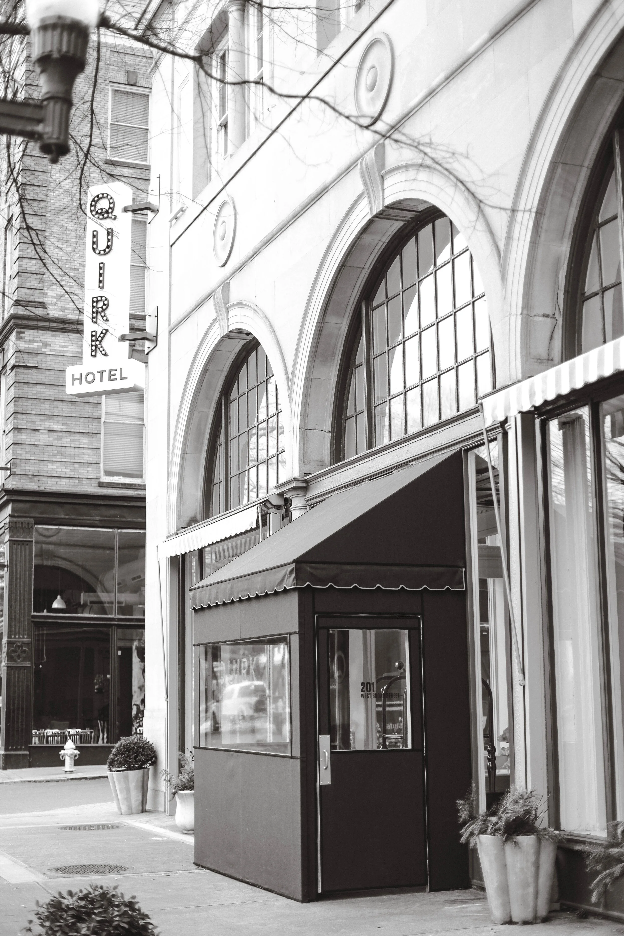 Black and white photo of a city street showing a hotel with a sign that reads 'QURK HOTEL' and a small storefront with a window and potted plants outside.