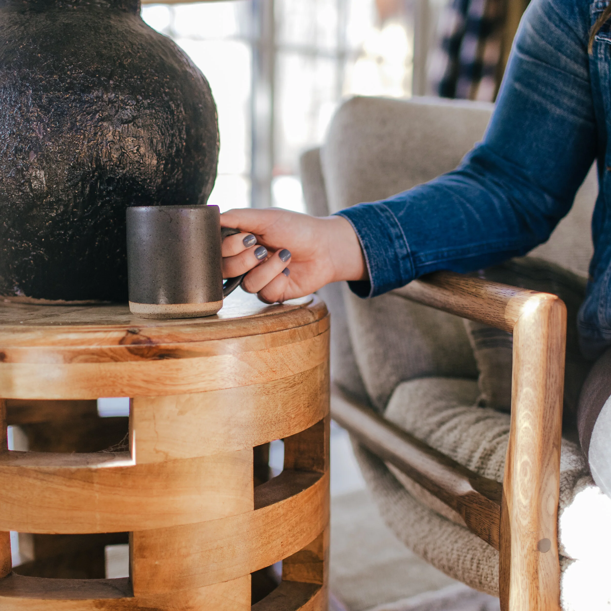 A person in a blue denim jacket sitting on a wooden chair, reaching to pick up a black mug on a round wooden table, with a large, dark textured object and a light-colored sofa in the background.