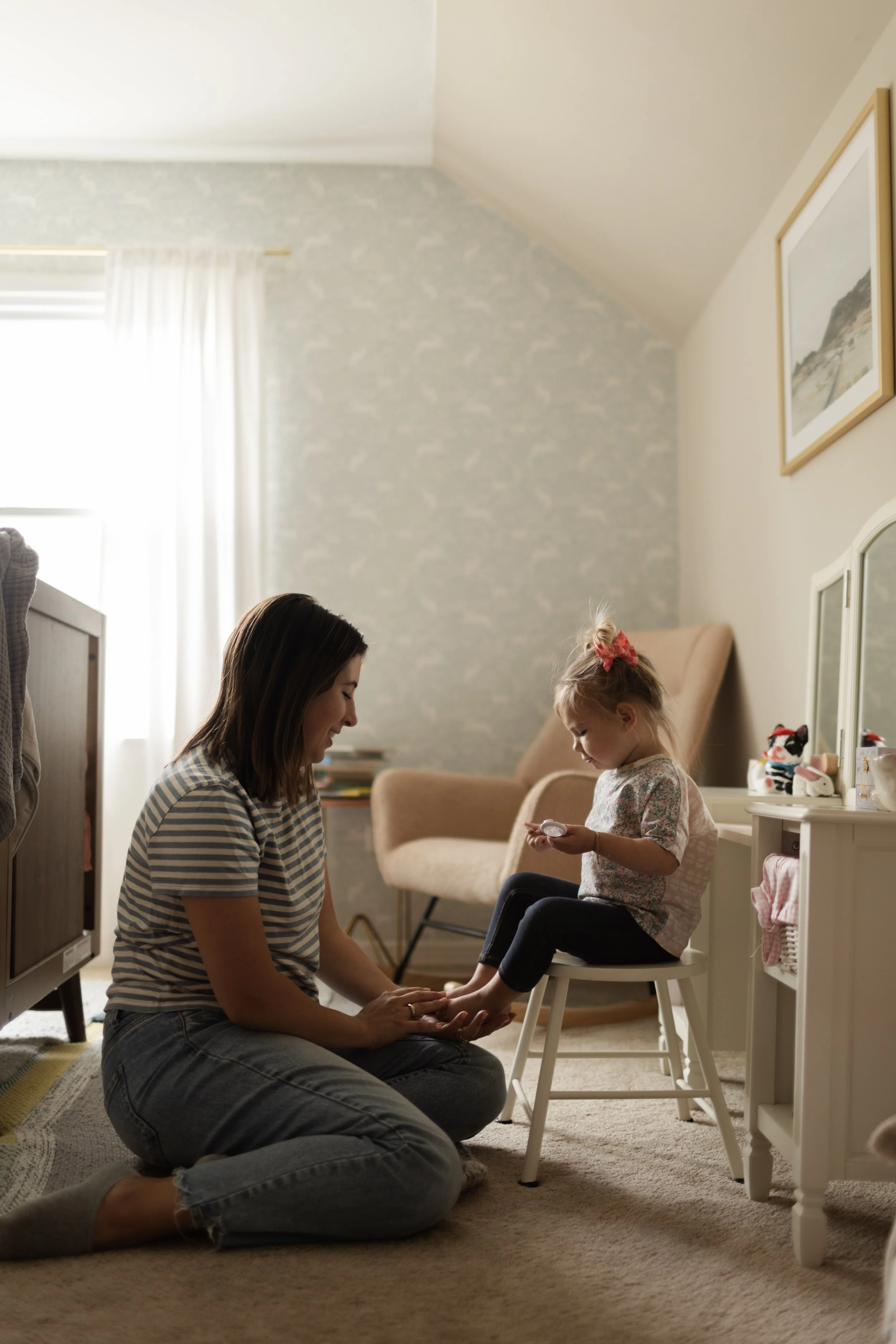 A woman and a young girl sitting on the floor in a cozy bedroom, with the woman holding the girl's feet in her hands and the girl looking at a small container, opposite a white dresser with toys, in natural light from a window.