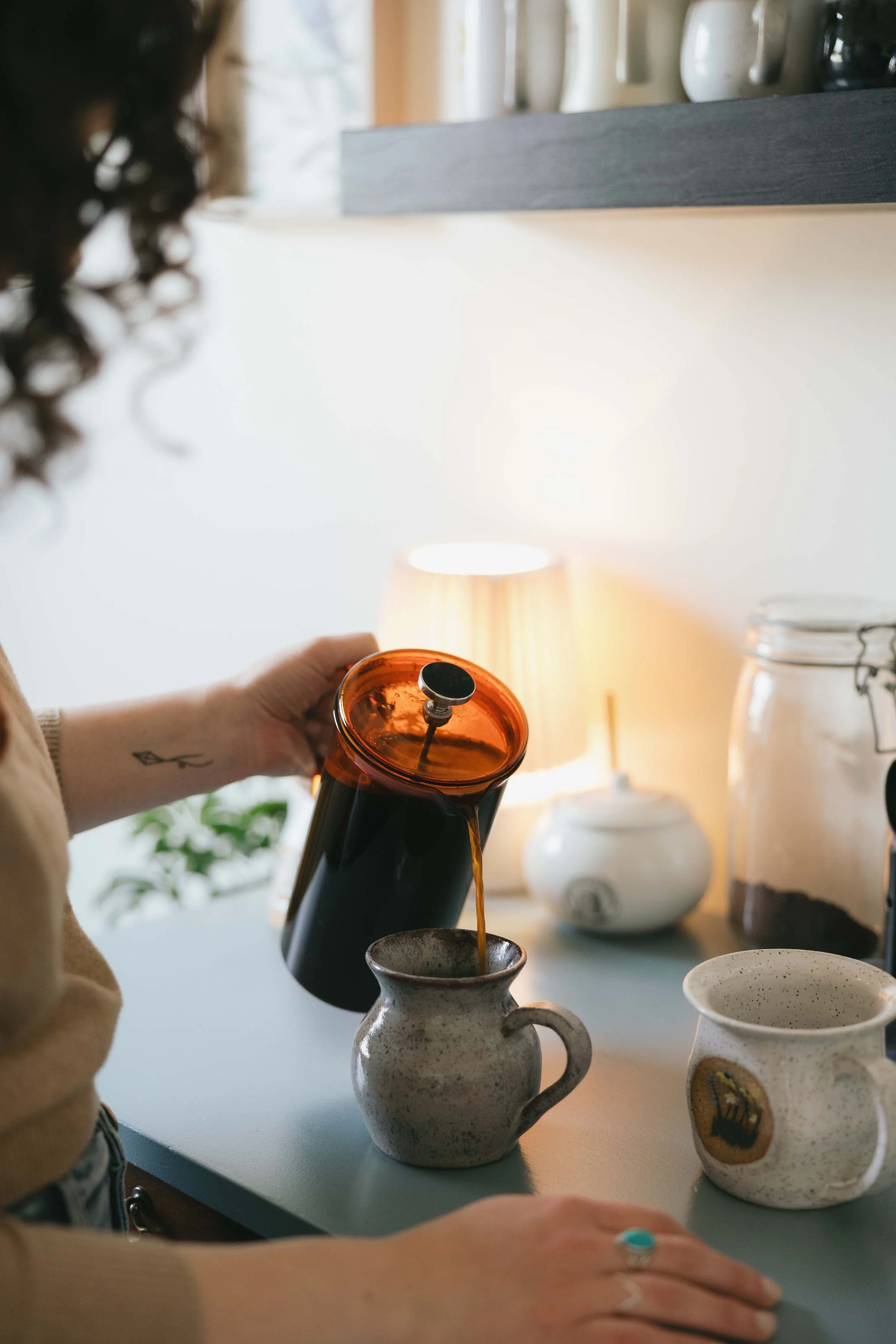 Person pouring tea from a brown teapot into a ceramic mug on a light-colored table in a cozy, well-lit room with decorative items and a lamp in the background.