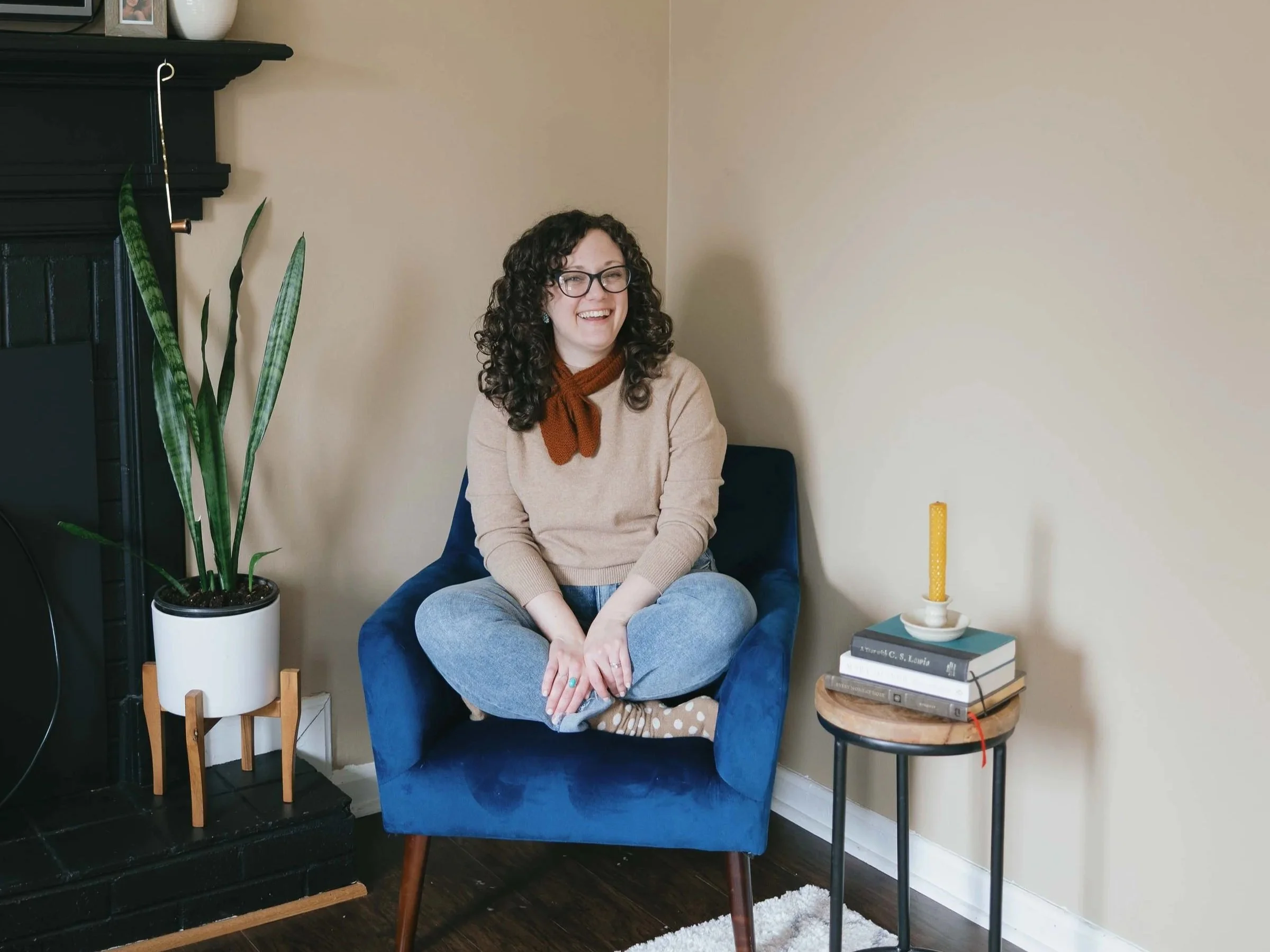 A woman with curly dark hair, glasses, and a smile, sitting cross-legged in a blue armchair in a living room. She is wearing a beige sweater, a rust-colored scarf, and polka-dot socks. To her left is a large potted plant with tall, green leaves. To her right, there is a stack of books on a small round wooden table with a yellow candle on top. Behind her, there's a beige wall, and a black fireplace mantel with framed photos and decorative items. The floor is dark wood with a white rug partially visible.