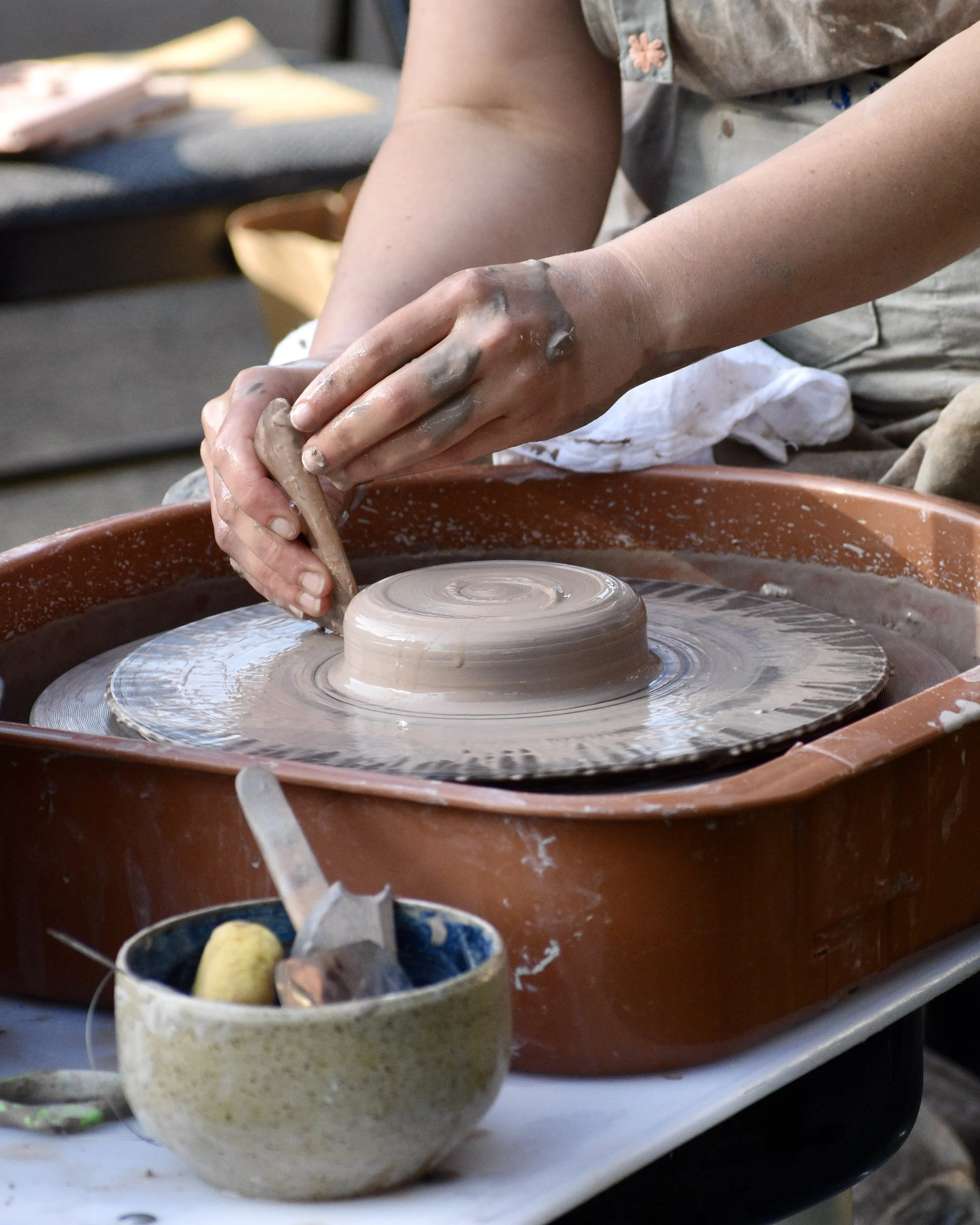 Person shaping clay on a spinning pottery wheel, with pottery tools and a cup of water in the foreground.