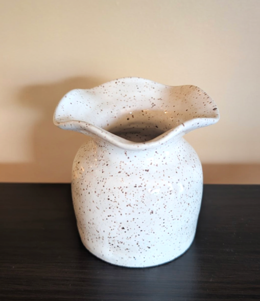 White ceramic vase with speckled brown pattern on a black surface against a beige wall.