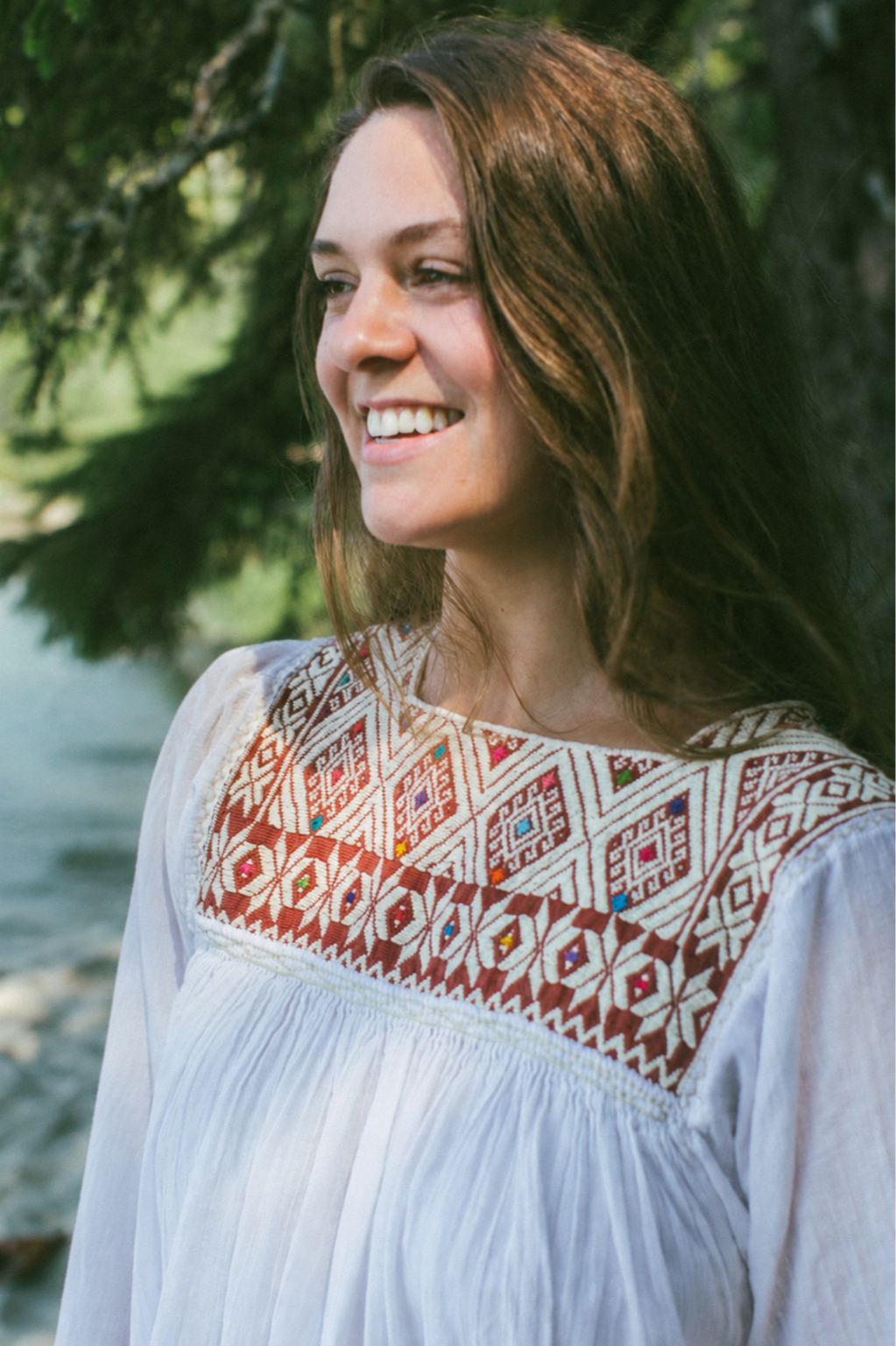 A woman with shoulder-length brown hair smiling outdoors, wearing a white dress with colorful embroidered patterns on the chest, with greenery in the background.