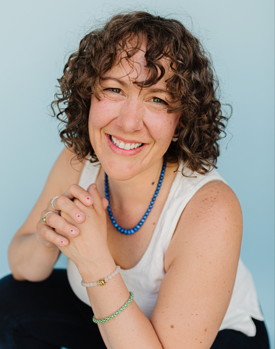 A woman with short, curly brown hair and light skin is smiling at the camera. She is wearing a white sleeveless top, a blue beaded necklace, and bracelets on her left wrist. Her hands are clasped together near her chin, and she is sitting against a light blue background.