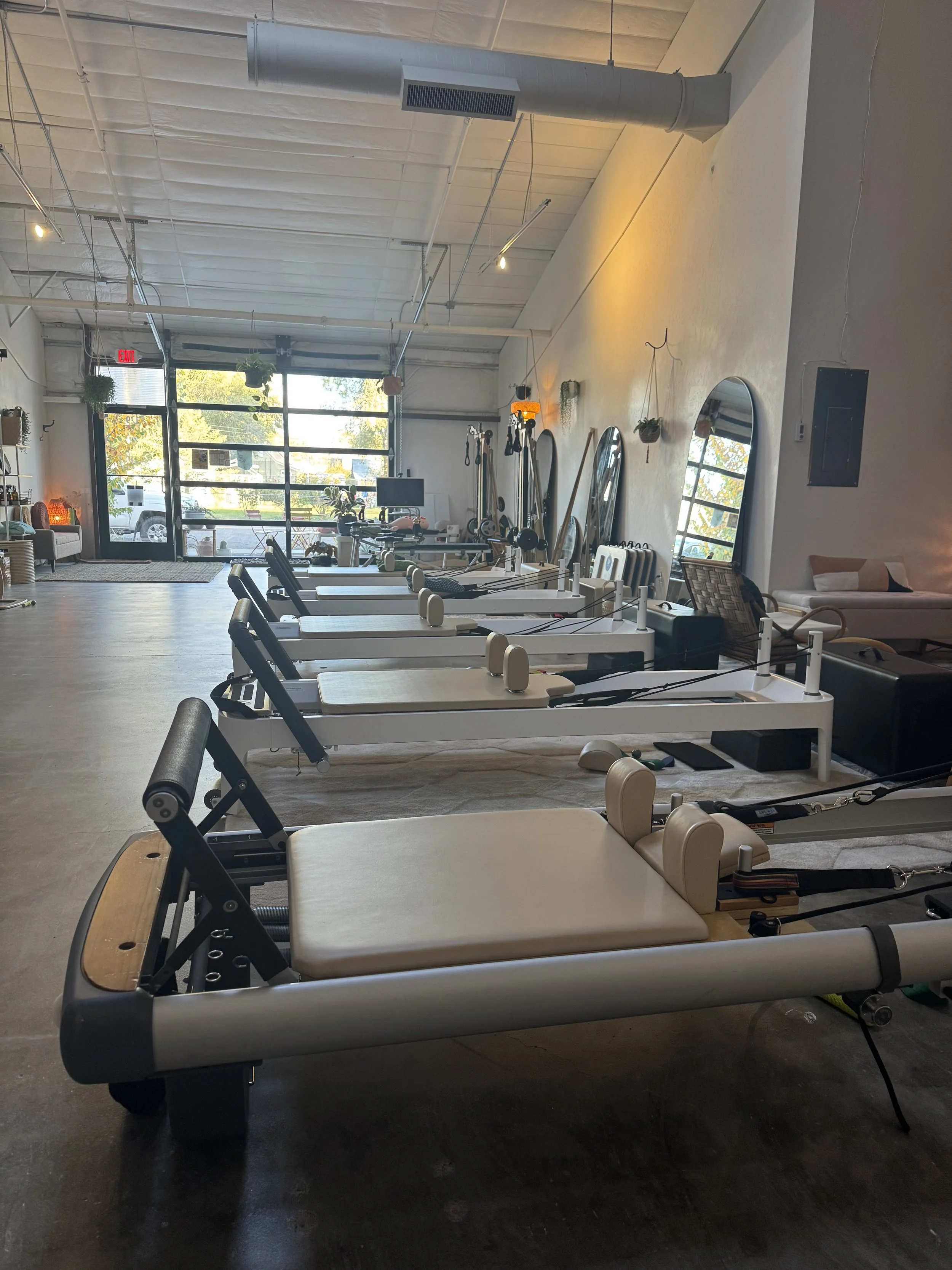 Interior of a Pilates studio with reformer Pilates machines lined up, mirrors on the wall, and a large glass front door letting in natural light.