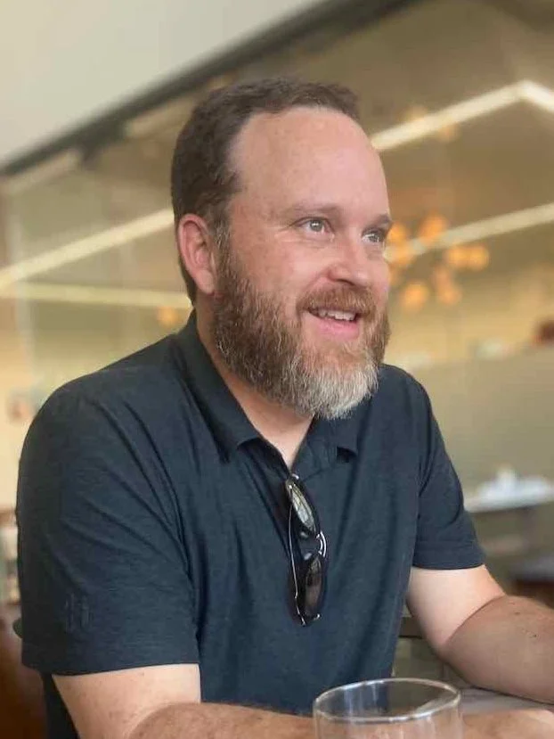 A smiling man with a beard and short hair wearing a dark polo shirt, sitting at a table with glasses in front of him in a well-lit indoor setting, possibly a restaurant or cafe.