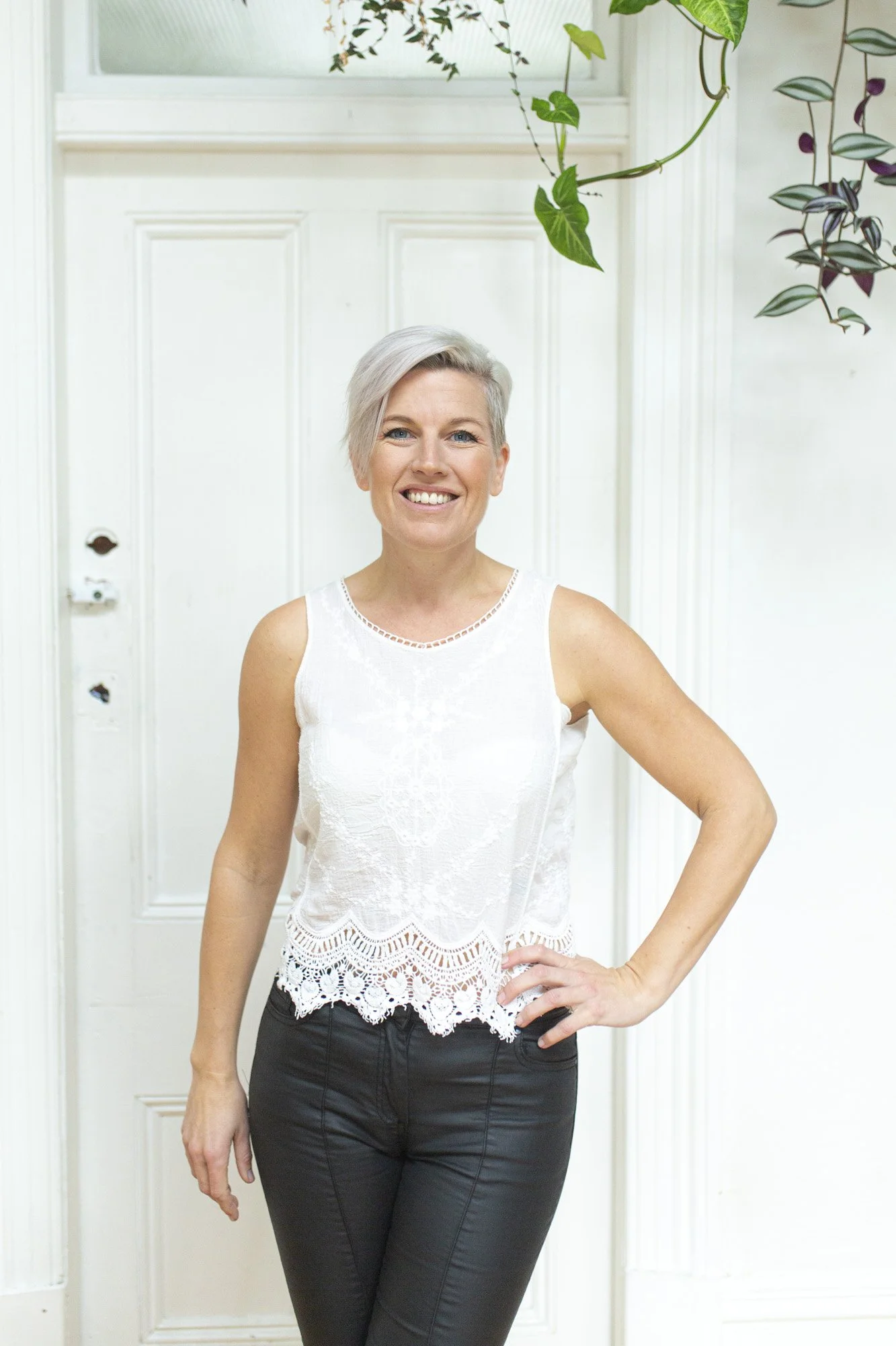A woman with short blonde hair smiling and standing with one hand on her hip, wearing a sleeveless white lace top and black pants, in front of a white door and plants.