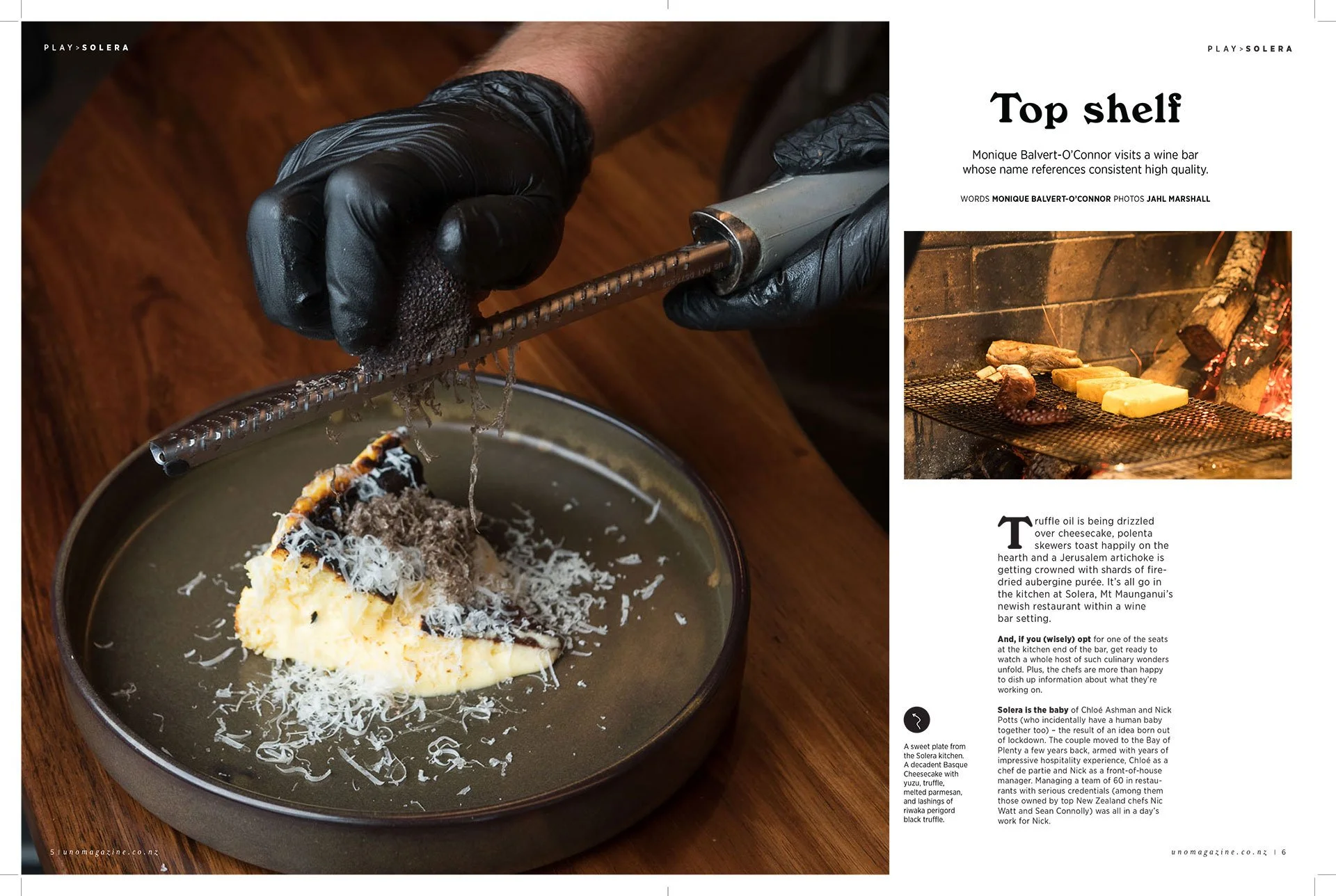 A person wearing black gloves uses tongs to handle a piece of food over a metal bowl containing mashed potatoes and grated cheese. In the background, there is a fireplace with hot cheese melting on a piece of bread on a grill.