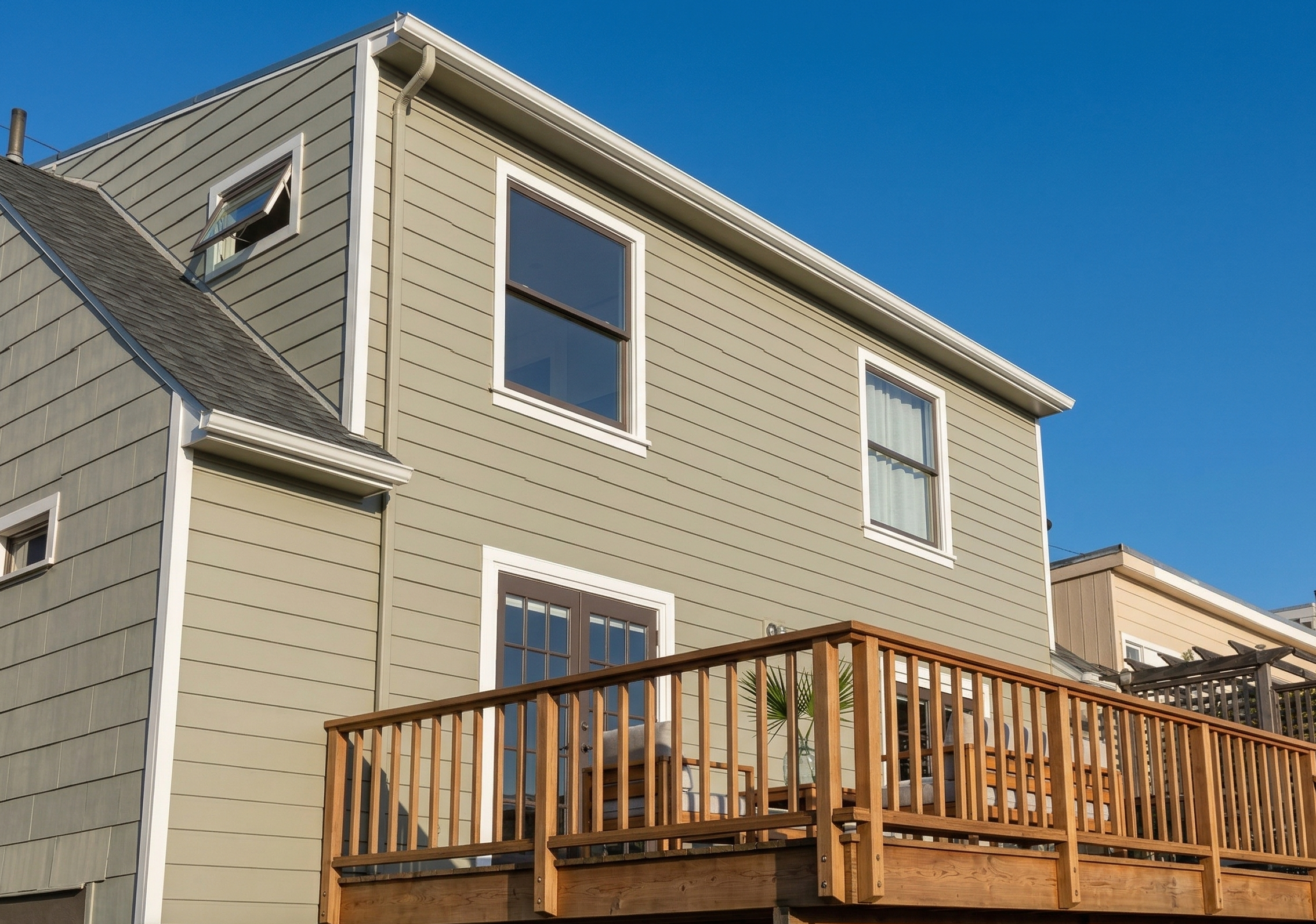 Exterior of a beige house with multiple windows and a wooden deck with outdoor furniture, under a clear blue sky.