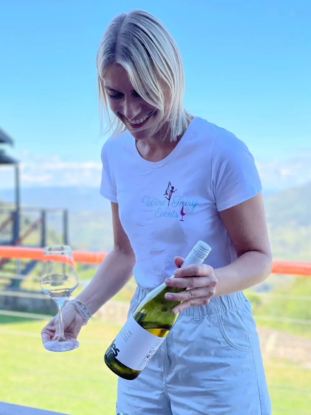 Kat, founder of Wine Fairy Events, smiling while pouring wine in her Wine Fairy shirt