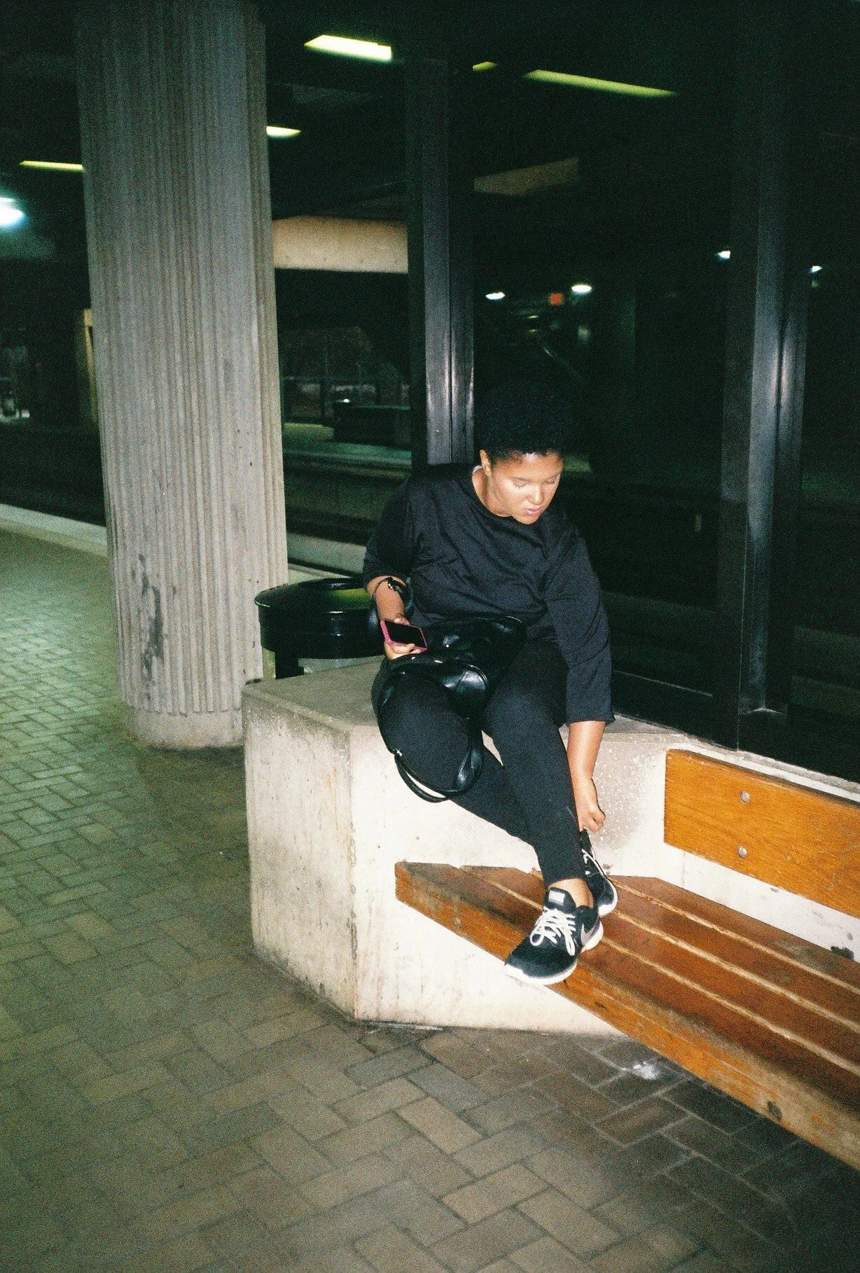 A young woman sitting on a bench at a bus stop at night, looking down and adjusting her shoe. She holds a smartphone in one hand and has a black handbag next to her.