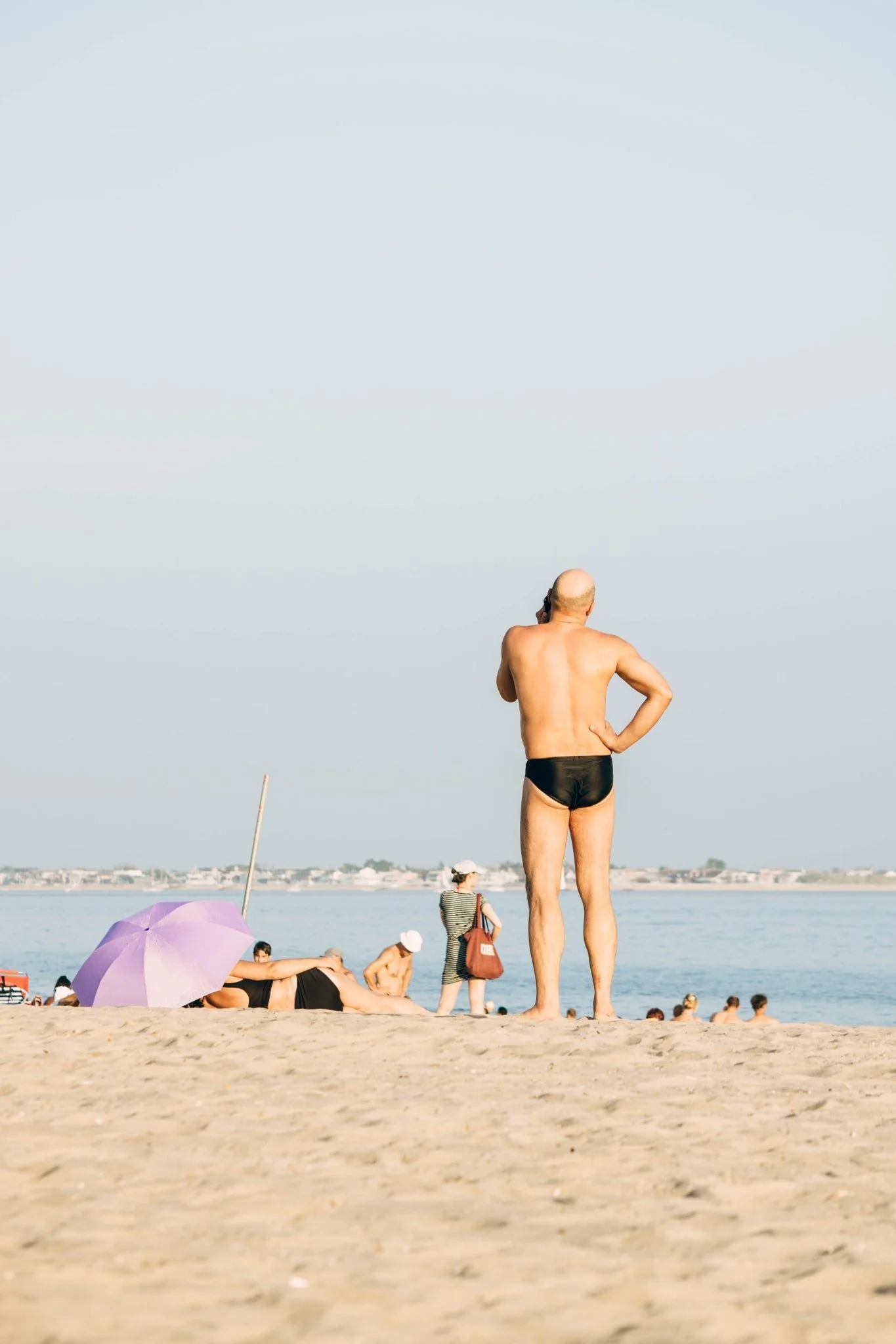 A man standing on a sandy beach looking out at the water, with other people and umbrellas in the background.