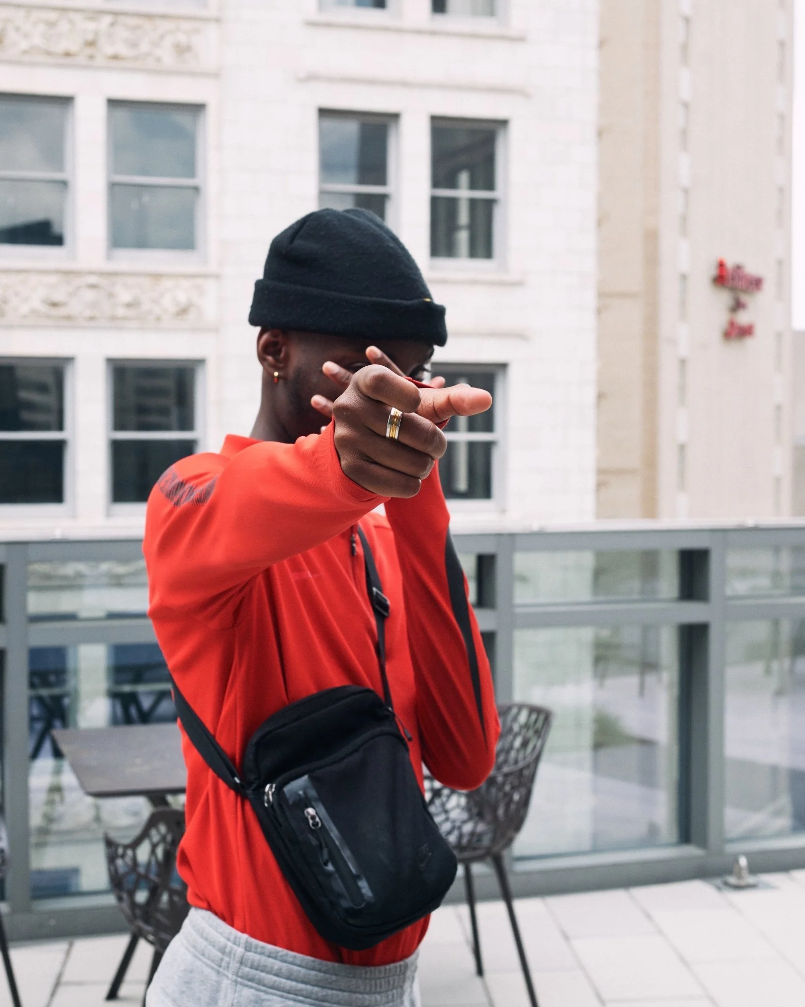 A man wearing a black beanie, a red jacket, and grey sweatpants, pointing at the camera, standing on a balcony with a black crossbody bag.