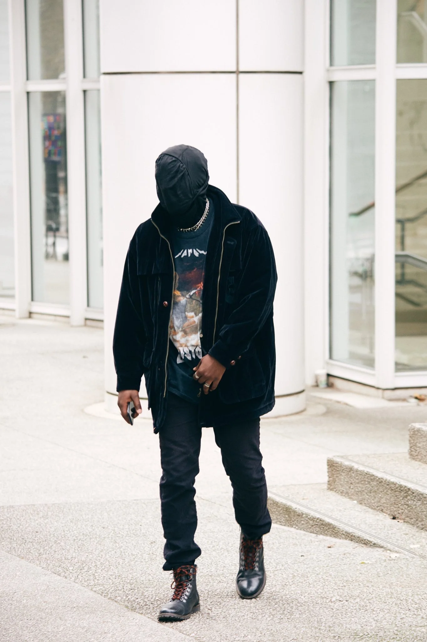 A person dressed in dark clothing, including a cap, jacket, and boots, walking outside near modern glass and metal building.