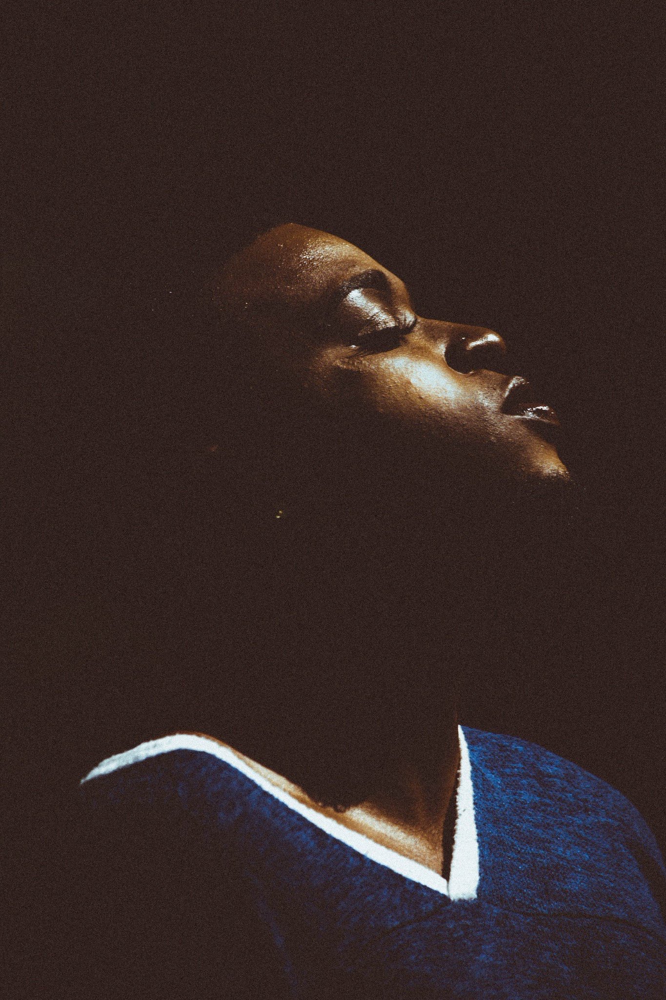 Close-up of a person with dark skin and short hair, highlighted by a light, against a dark background.
