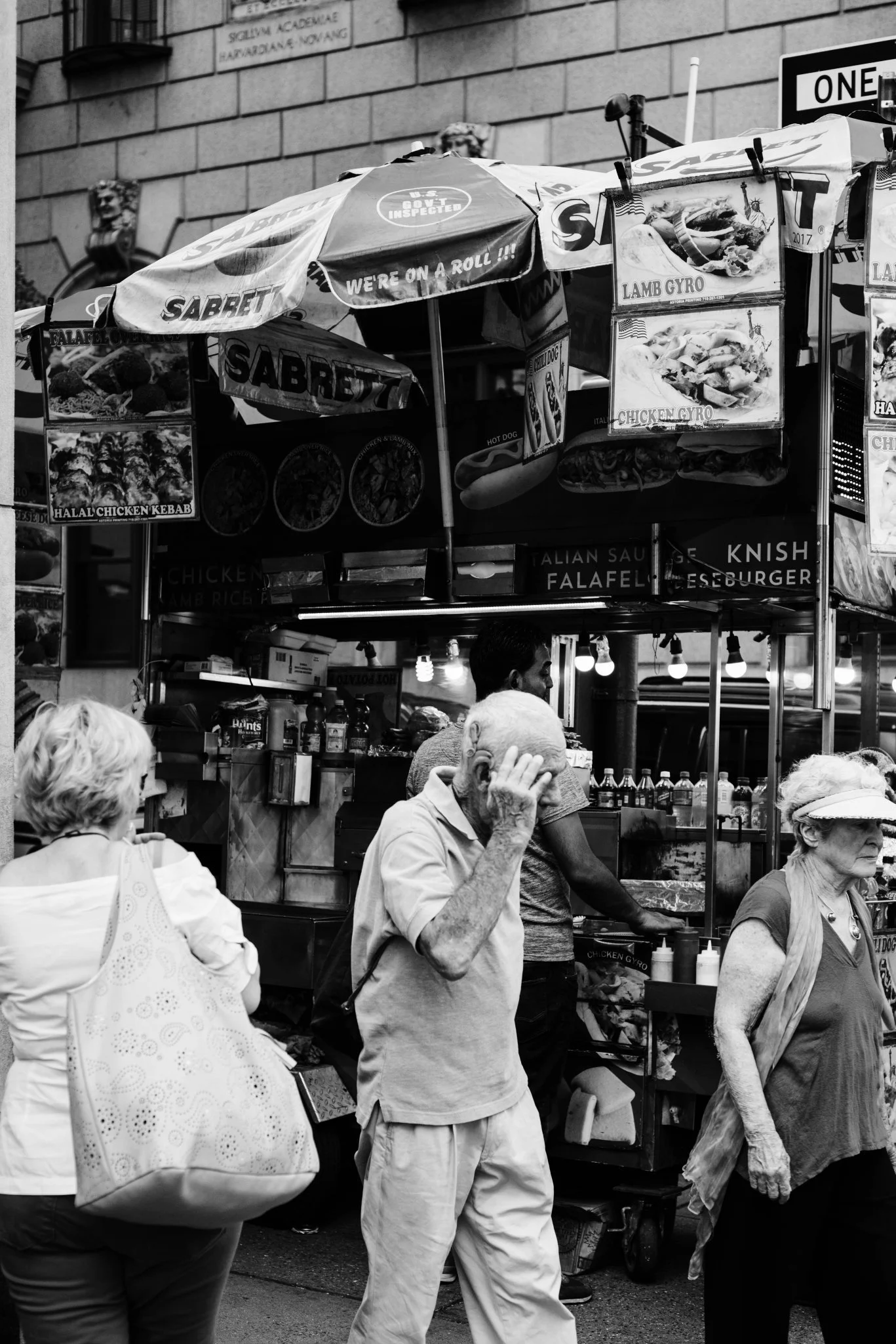 A busy street food cart selling lamb and chicken gyros, with several people walking by, including an elderly man covering his face and two women, one with a large patterned bag. The cart has signs and umbrellas advertising the food items.