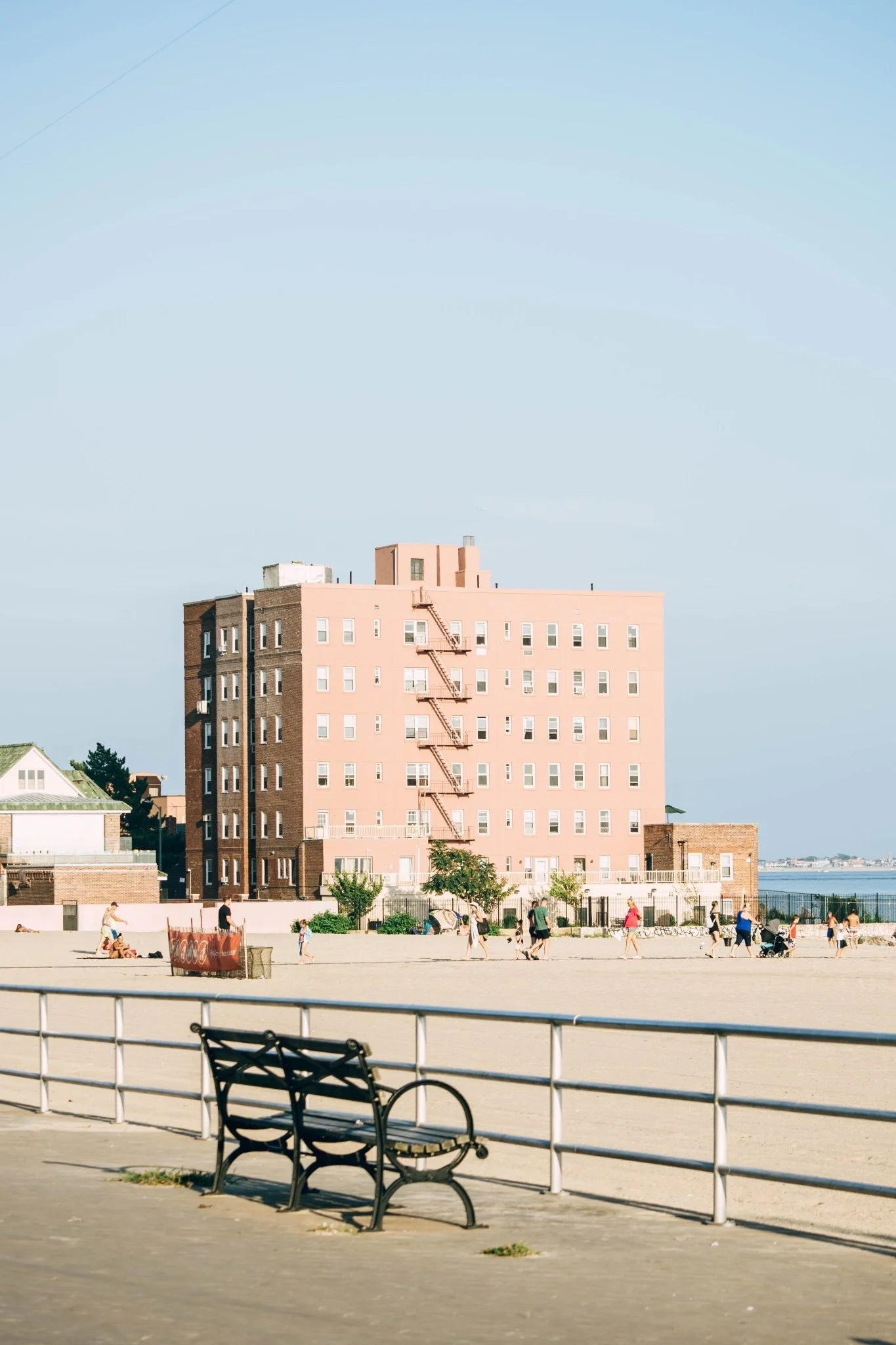 A pink apartment building near a beach with people walking and a bench in the foreground.