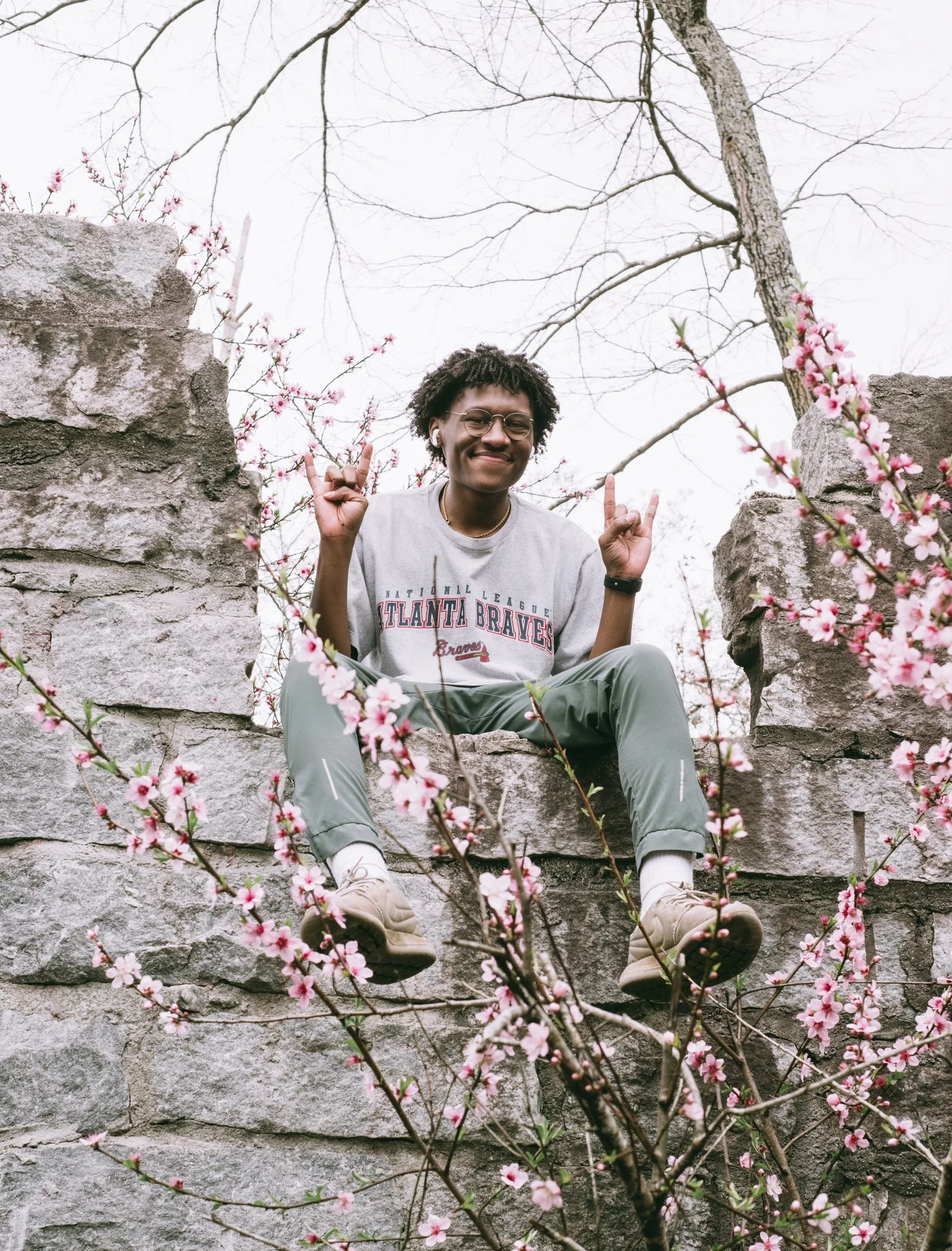 A young person sitting on a stone wall among pink flowering branches, smiling and making a peace sign with both hands. They are wearing glasses, a gray Atlanta Braves t-shirt, gray pants, and light-colored sneakers.