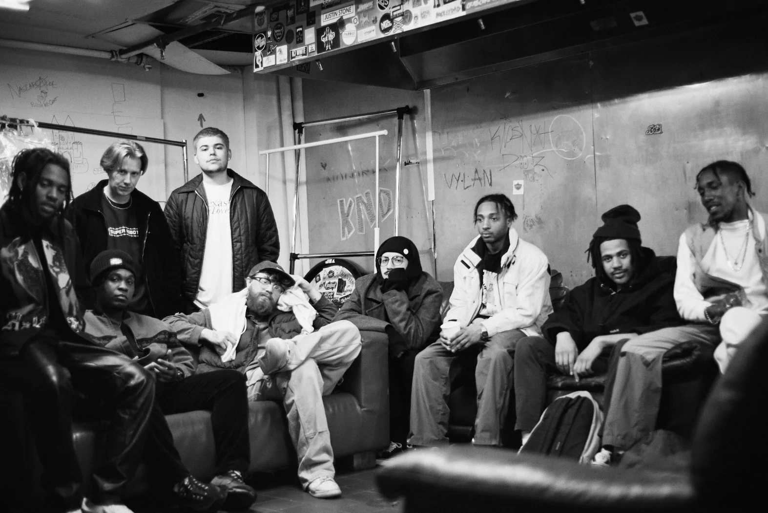 A black-and-white photo of nine young men in a room with graffiti-covered walls and various stickers. Some are seated on a couch and chairs, while others stand. They wear casual clothing and appear relaxed.