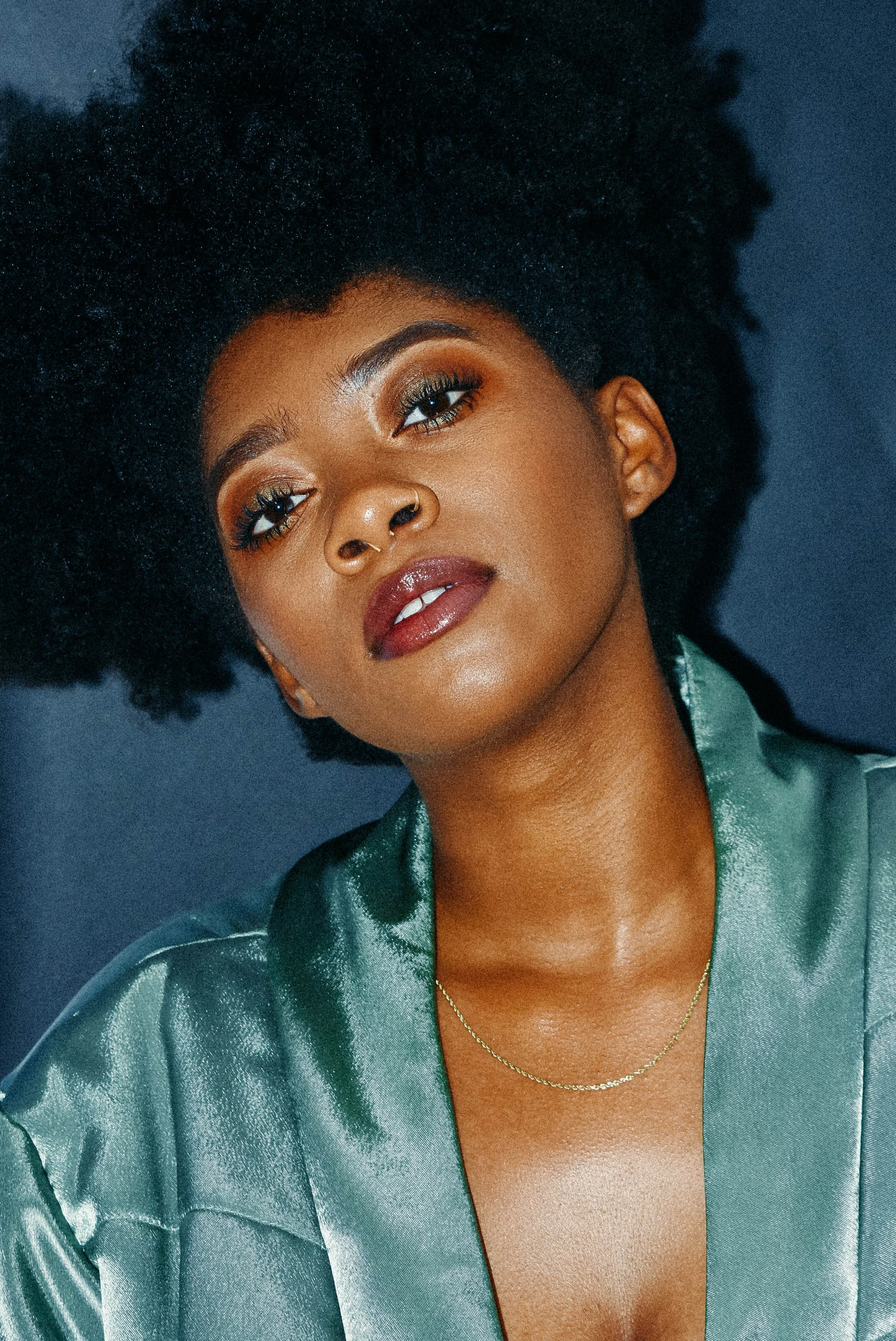 A close-up portrait of a woman with natural curly hair, wearing a green satin jacket and a delicate gold necklace, against a dark background.