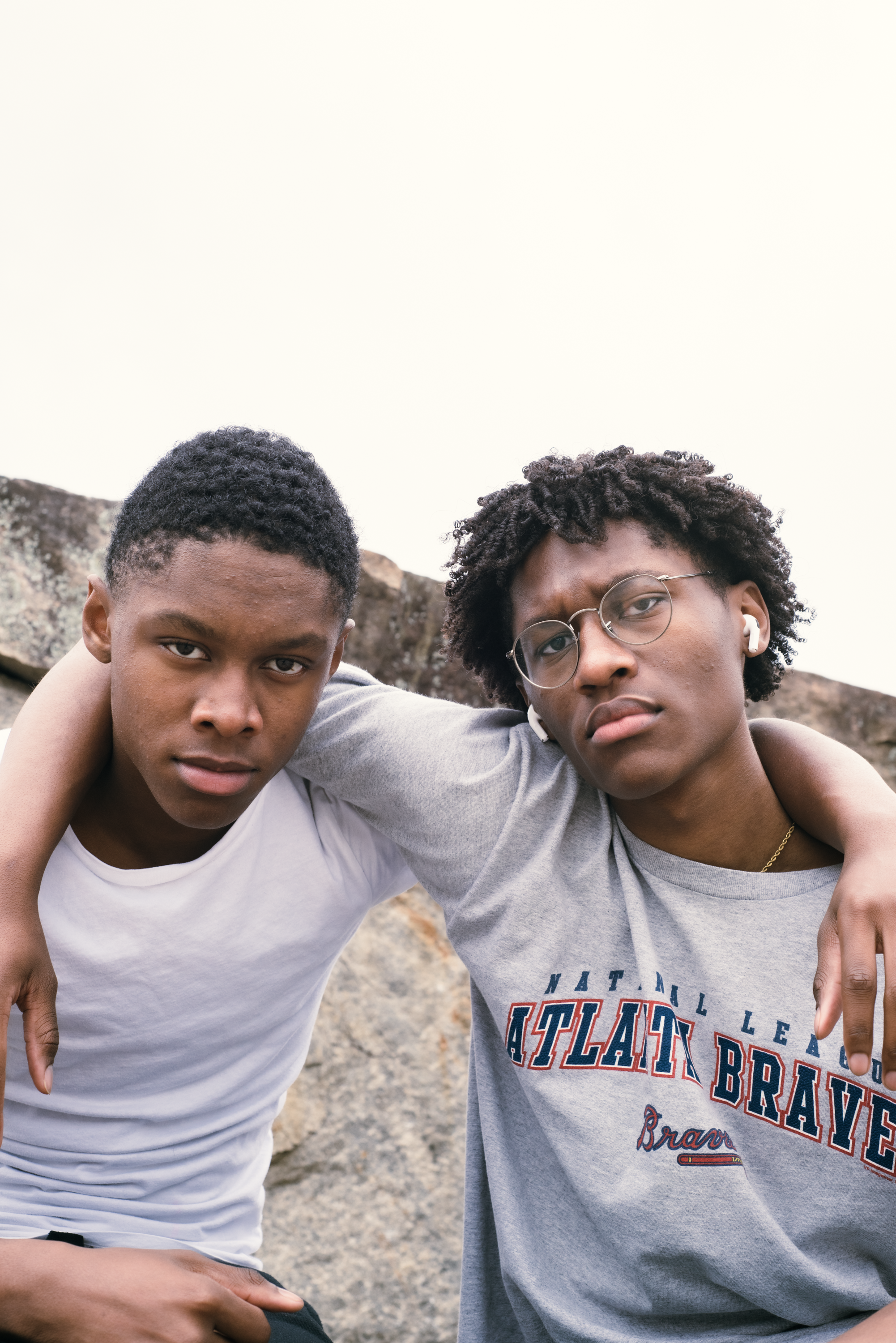 Two young men standing outdoors with their arms around each other, one wearing a white t-shirt and the other wearing glasses and a gray Atlanta Braves t-shirt, looking at the camera