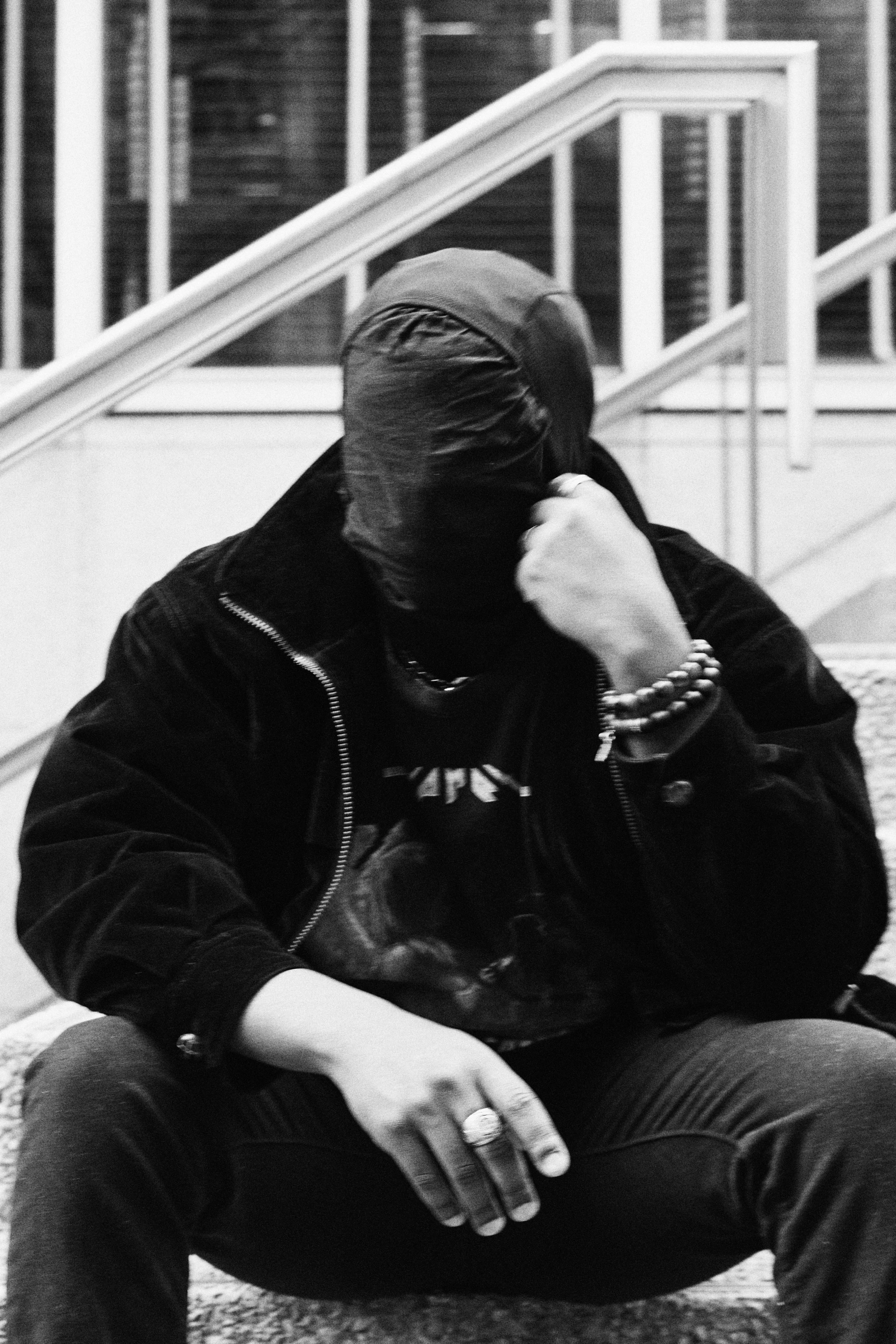 A person wearing a hooded jacket and a bandana over their face, sitting with hand on knee, in a black and white photo.
