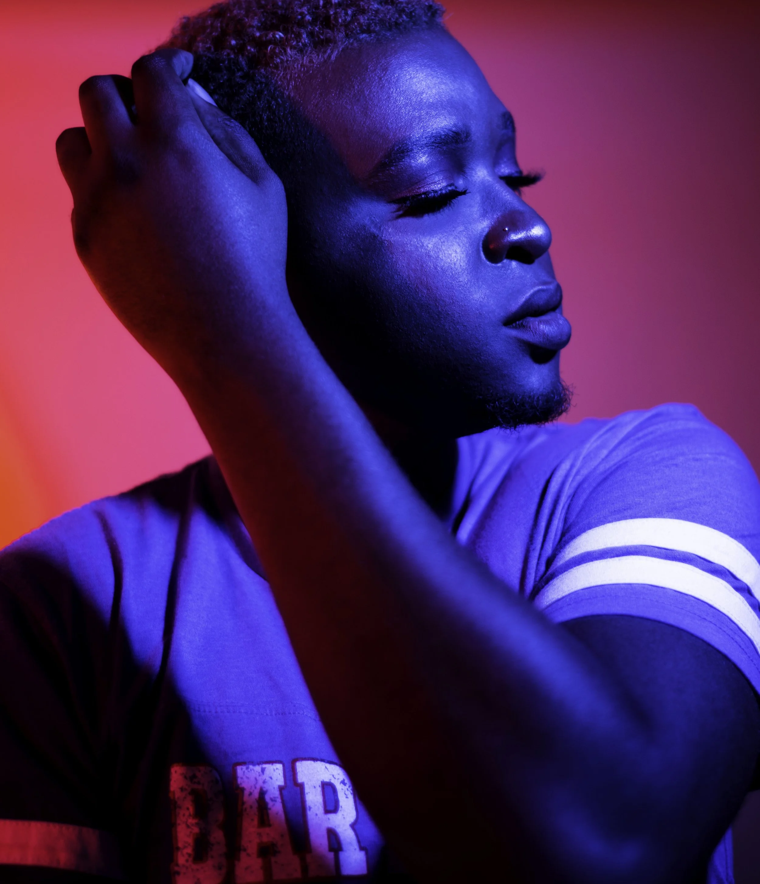 Close-up of a young man with dark skin and short curly hair, eyes closed, holding his head with one hand, against a pink and blue background, wearing a casual T-shirt.
