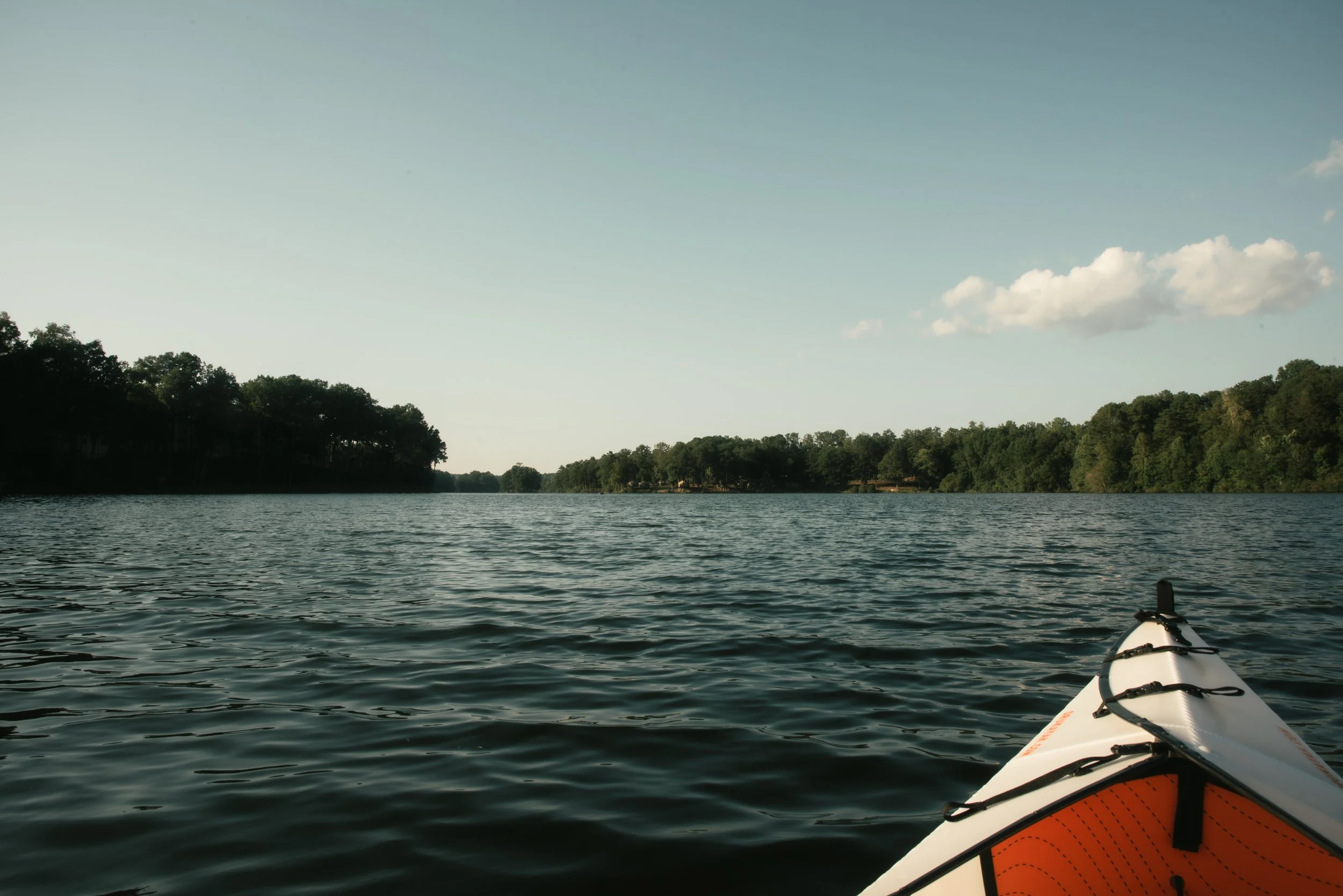 View from a kayak on a lake with water and distant tree-lined shoreline under a partly cloudy sky.