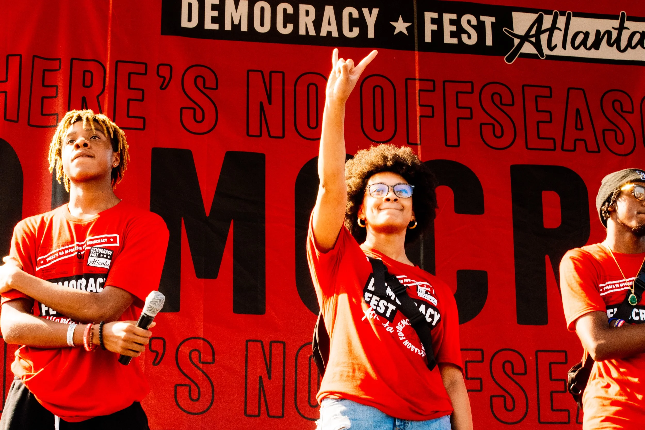 Three individuals standing on stage at the Democracy Fest in Atlanta, wearing red shirts, with a large red and black backdrop that reads 'Democracy Fest Atlanta' and 'Here's No Offseason' in the background. The person in the middle is smiling, wearing glasses, and making a peace sign with their right hand.