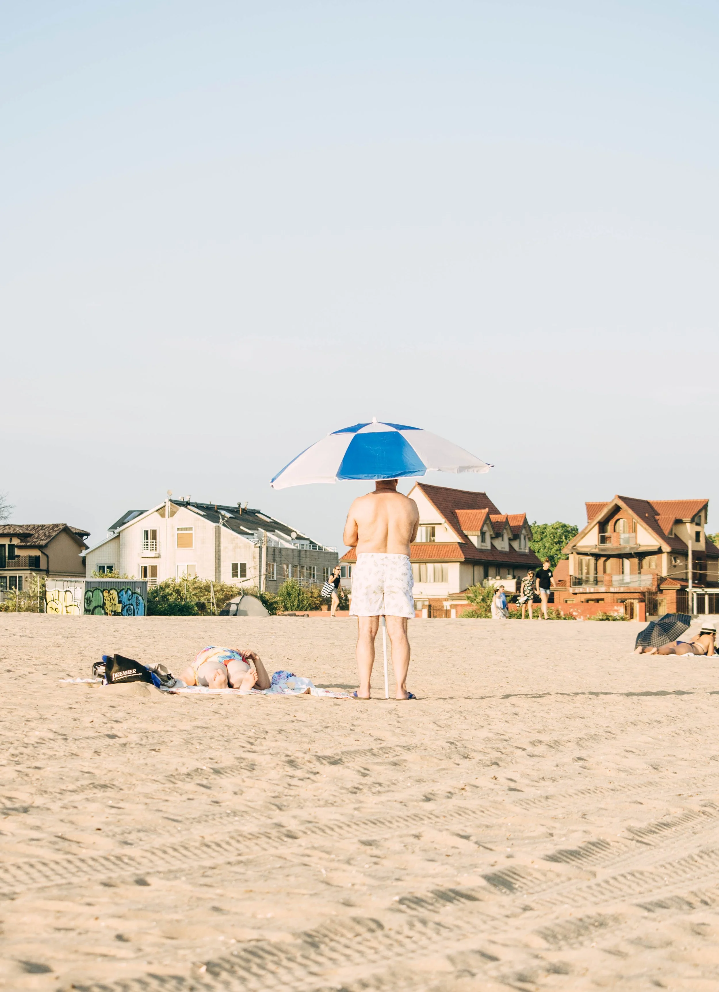 A man standing on a sandy beach with an umbrella, facing away from the camera. There are other beachgoers, houses, and a clear sky in the background.