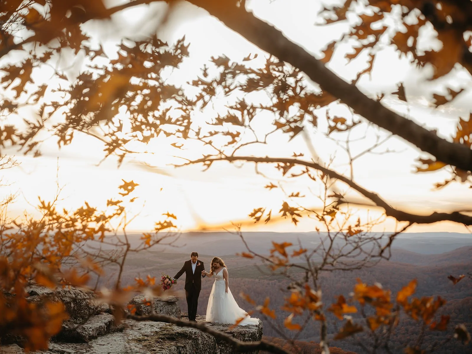 Bride and groom walking hand in hand on a mountaintop overlook in Arkansas at sunset, framed by autumn leaves, photographed by Arkansas wedding photographer Miles Witt Boyer.