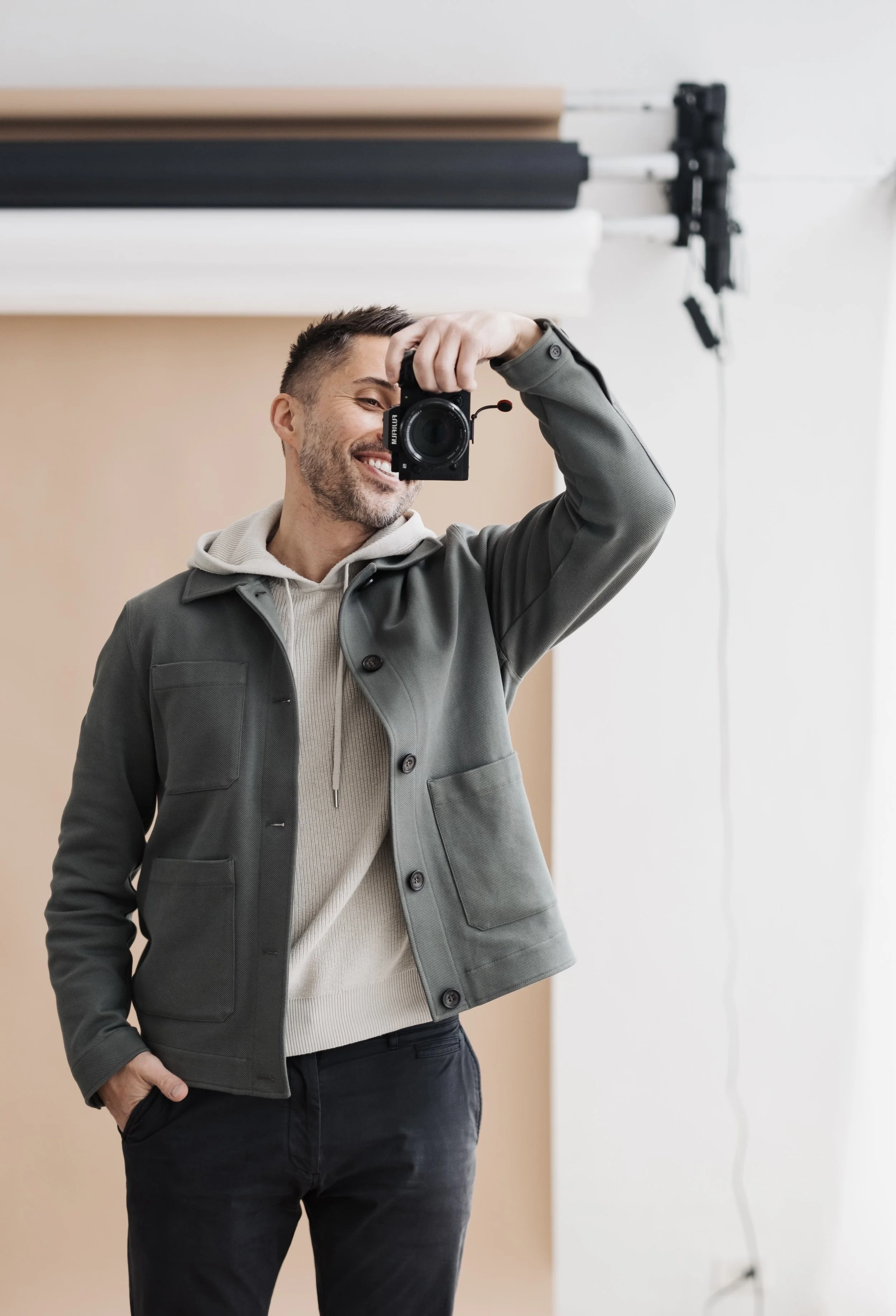 Man taking a selfie in a photography studio, holding a camera, smiling, with photography equipment in the background.