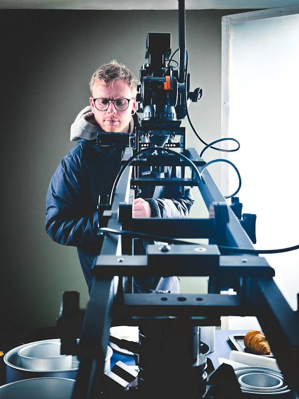 A man in glasses holds a camera jib with a camera on it over some pots on a table in a photography studio