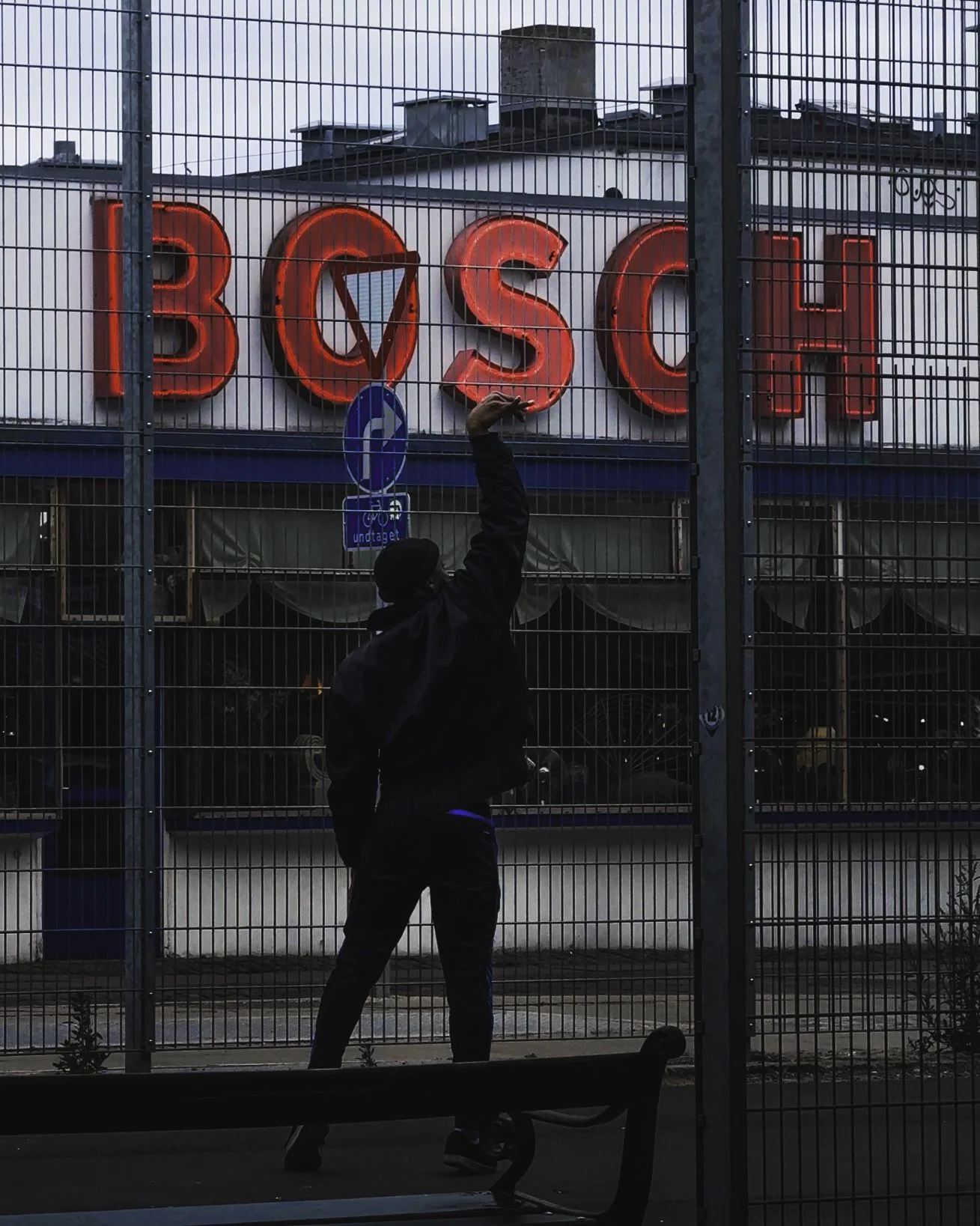 A person standing behind a metal fence, reaching up with one hand, in front of a building with a neon sign that reads 'BOSCH'.