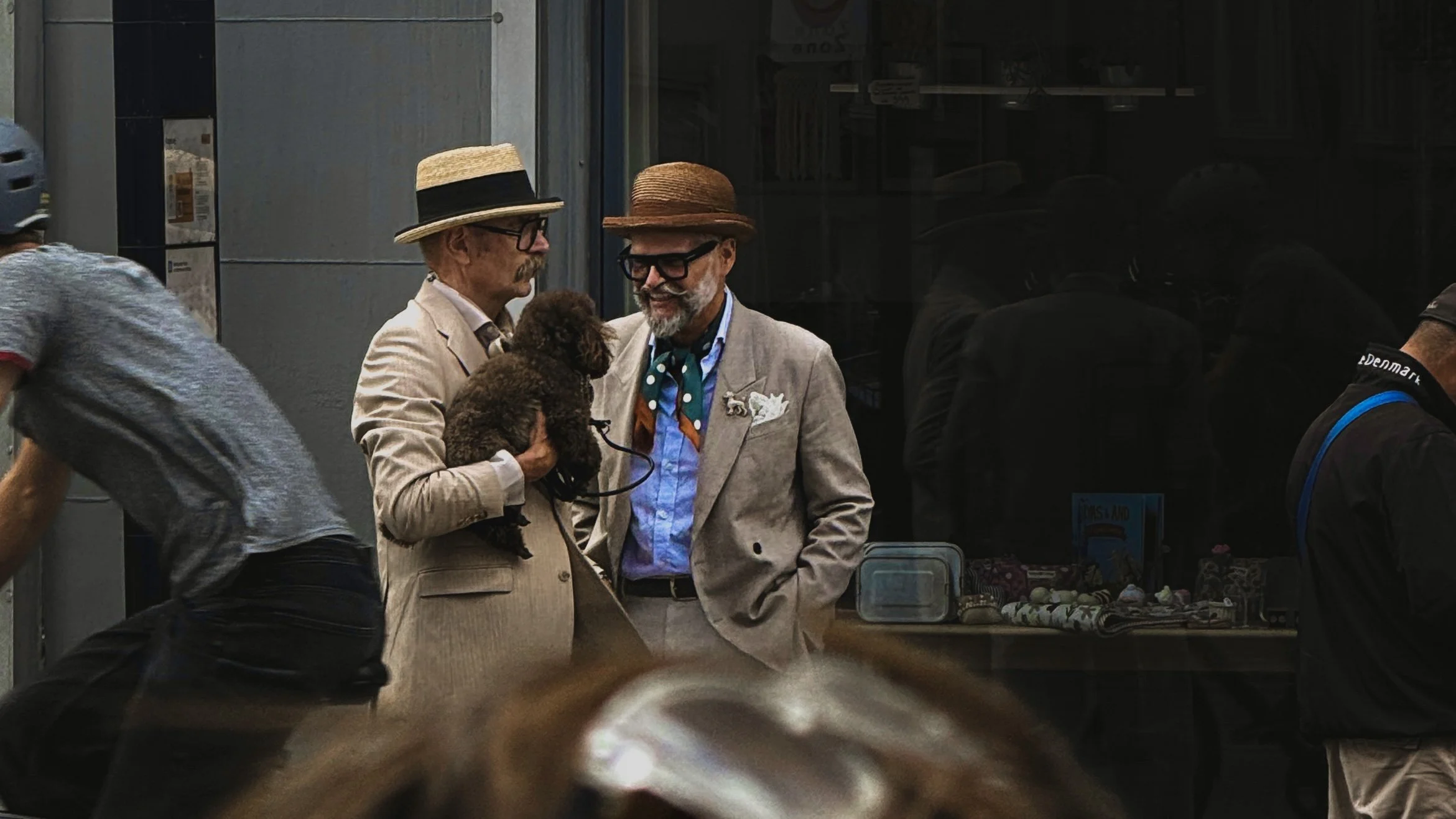 Two older men wearing straw hats and glasses, smiling and talking, one holding a small brown poodle dog. They are standing outside a storefront window.