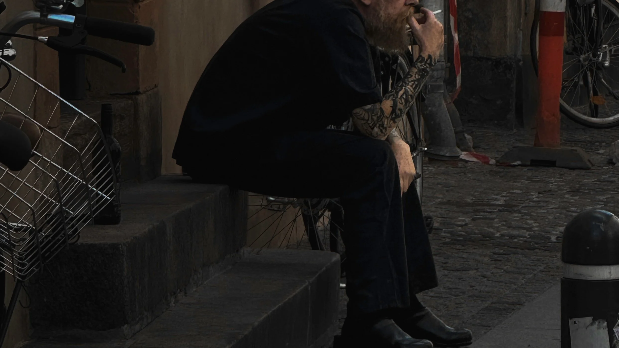 A man with tattoos on his arm sitting on a step, smoking a cigarette, next to a bicycle, with a red pole and a bicycle wheel in the background.