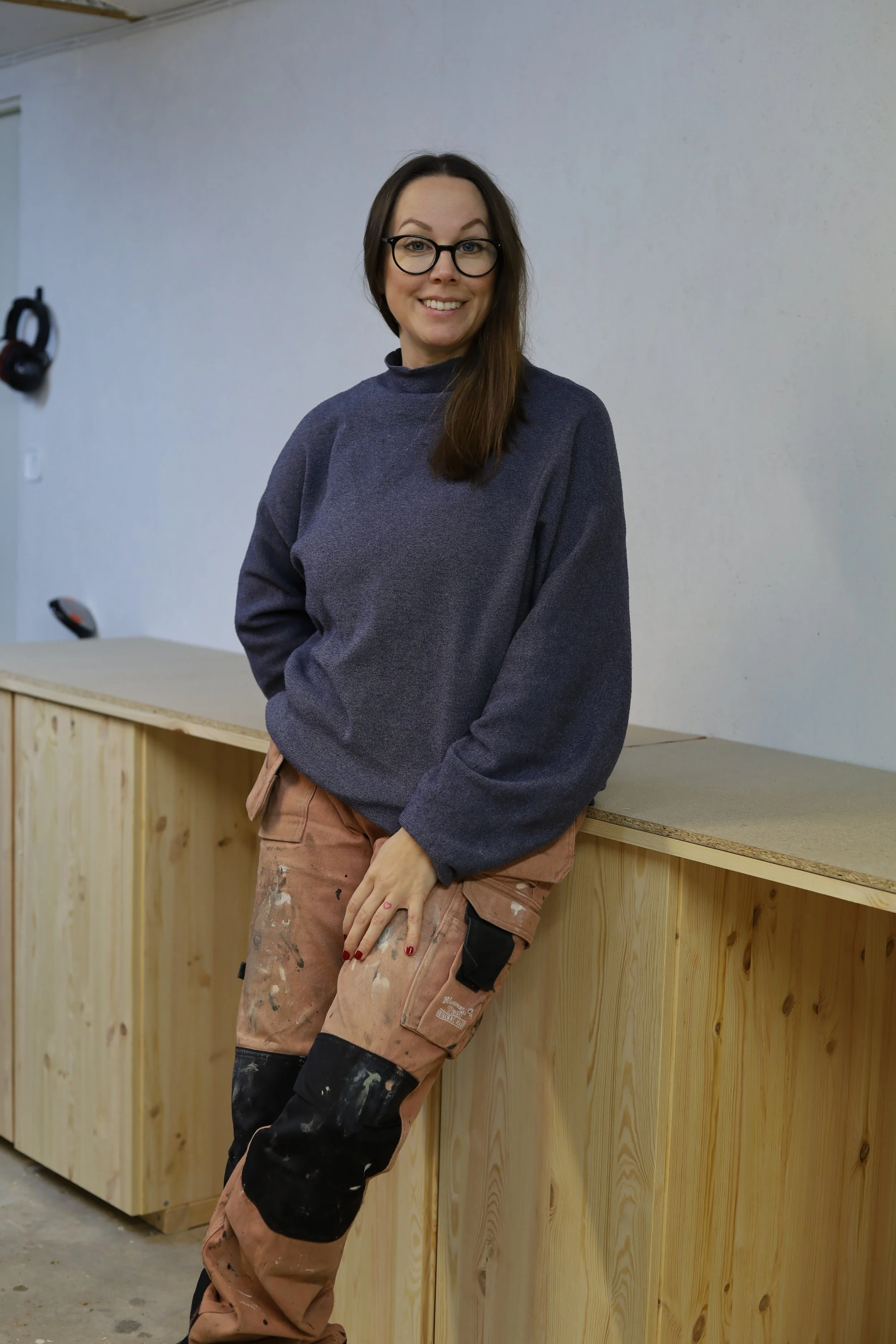 En kvinna med glasögon och långt brunt hår står lutad mot ett träbord i ett rum, klädd i en mörkblå tröja och arbetsbyxor.