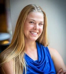A young woman with long blonde hair smiling, wearing a blue sleeveless top, in an indoor setting.