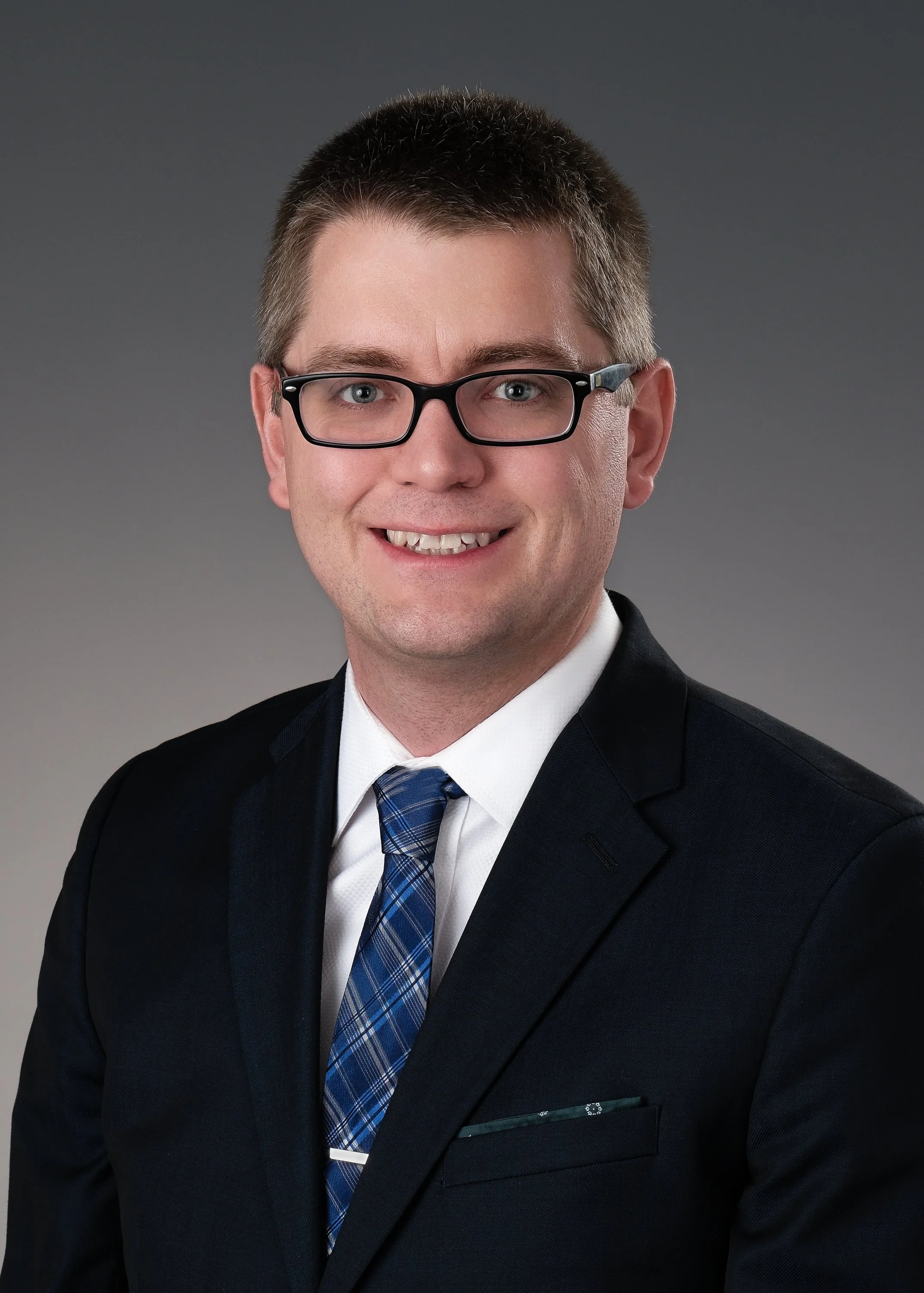 A professional headshot of a man in a dark suit, white shirt, and a blue patterned tie, wearing glasses, smiling against a gray gradient background.