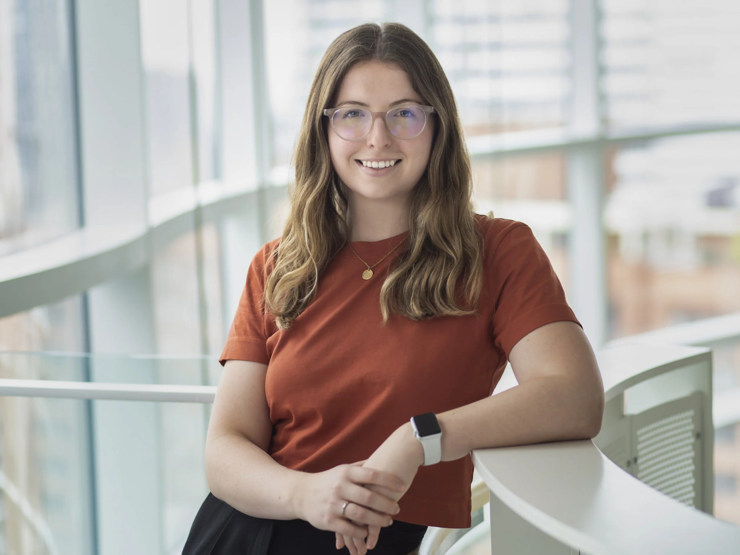 Young woman with glasses and long wavy hair smiling, wearing a rust-colored shirt and a smartwatch, standing in a modern office building with large windows.