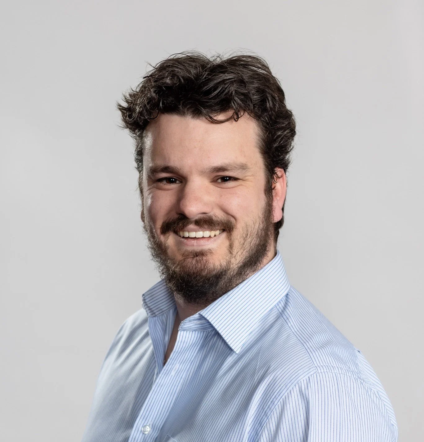 Headshot of a smiling man with dark, curly hair and a beard, wearing a light blue striped shirt against a plain light gray background.