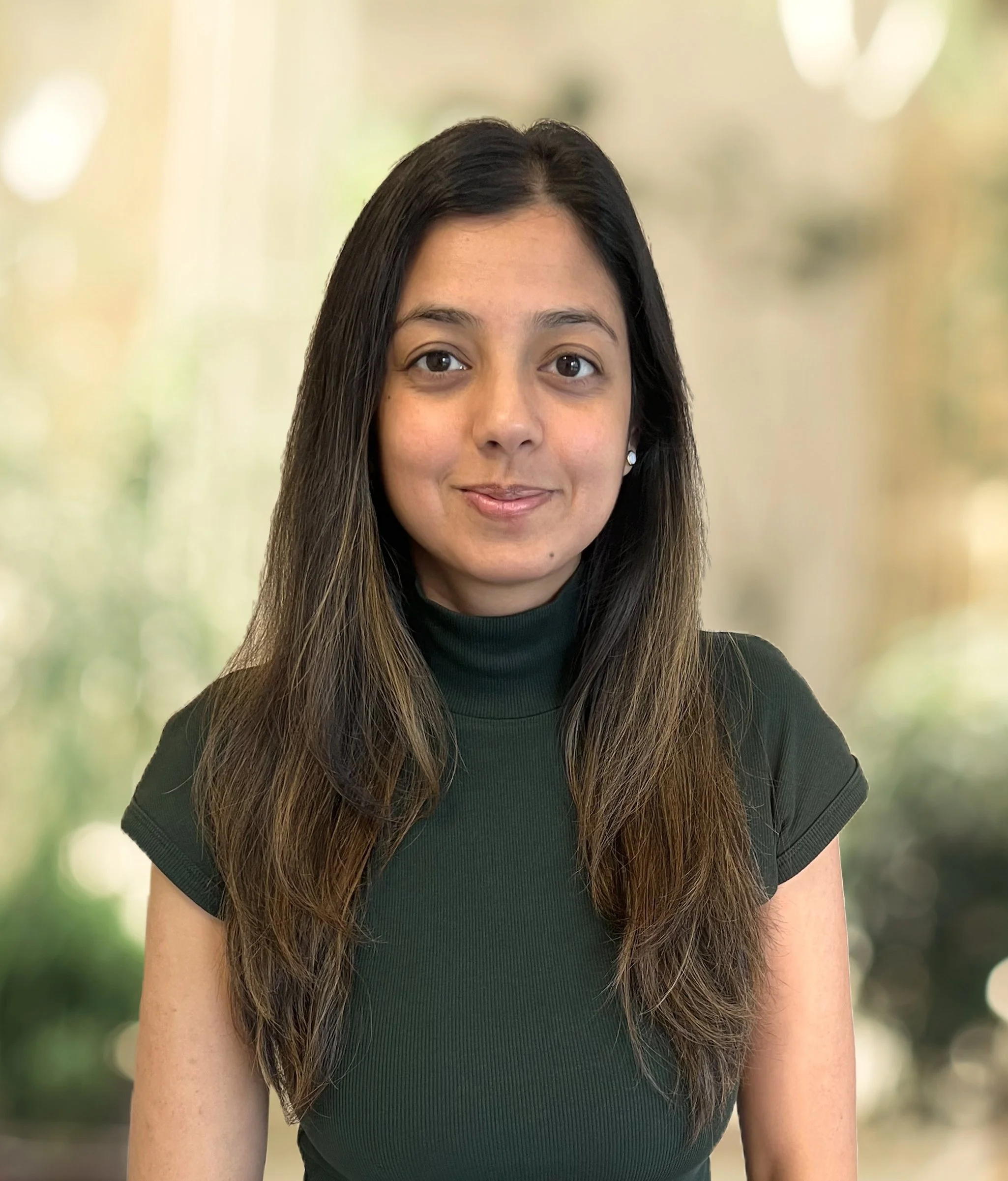 A young woman with long brown hair related to a blurred outdoor background, wearing a dark green turtleneck top and small earrings.