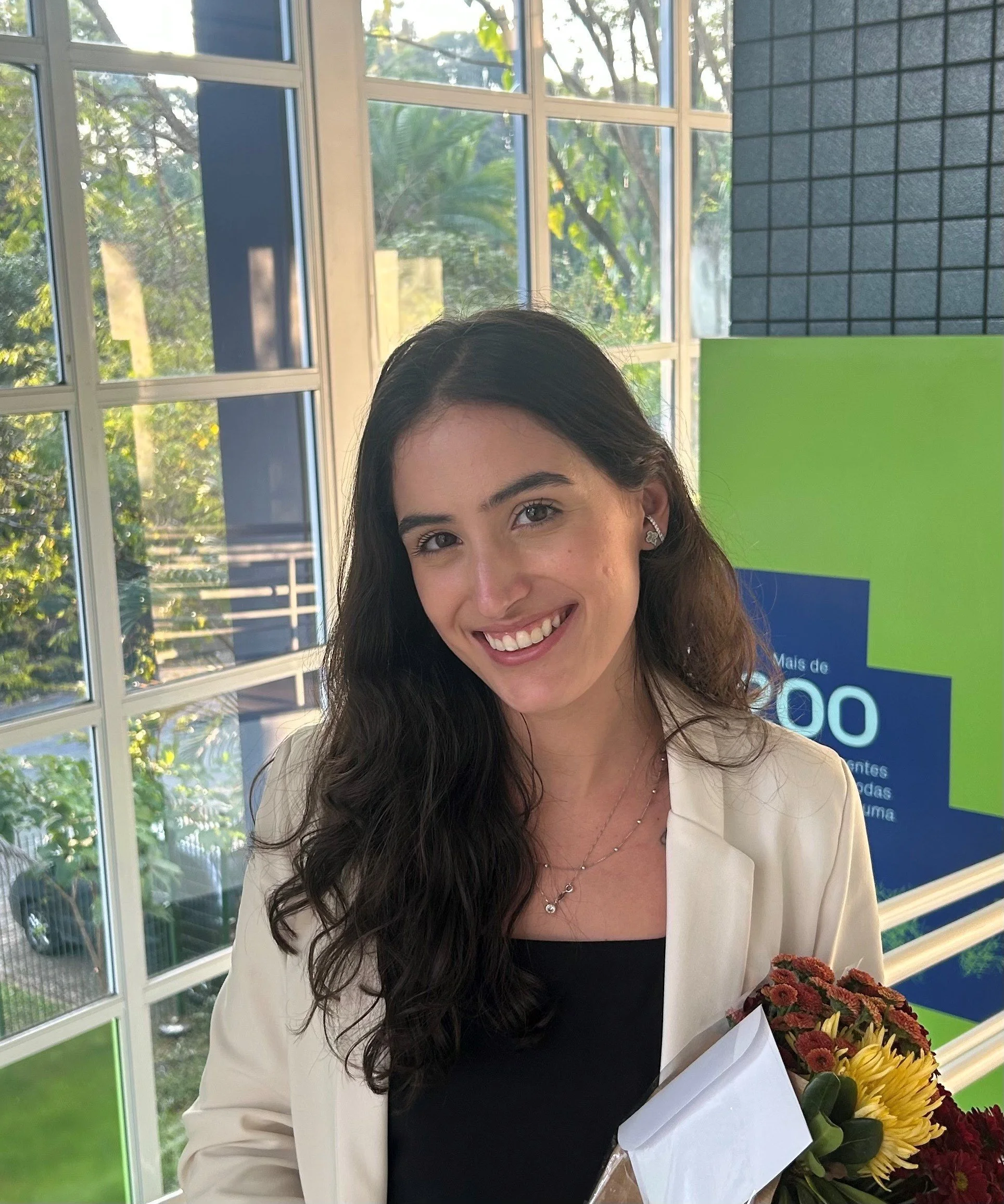 A woman with long dark hair wearing a white blazer and black shirt holding a bouquet of flowers, standing indoors near large windows with a view of trees outside.