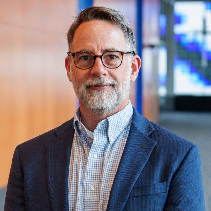 Professional headshot of a man wearing glasses, a dark blazer, and a blue checked collared shirt against a blue gradient background.