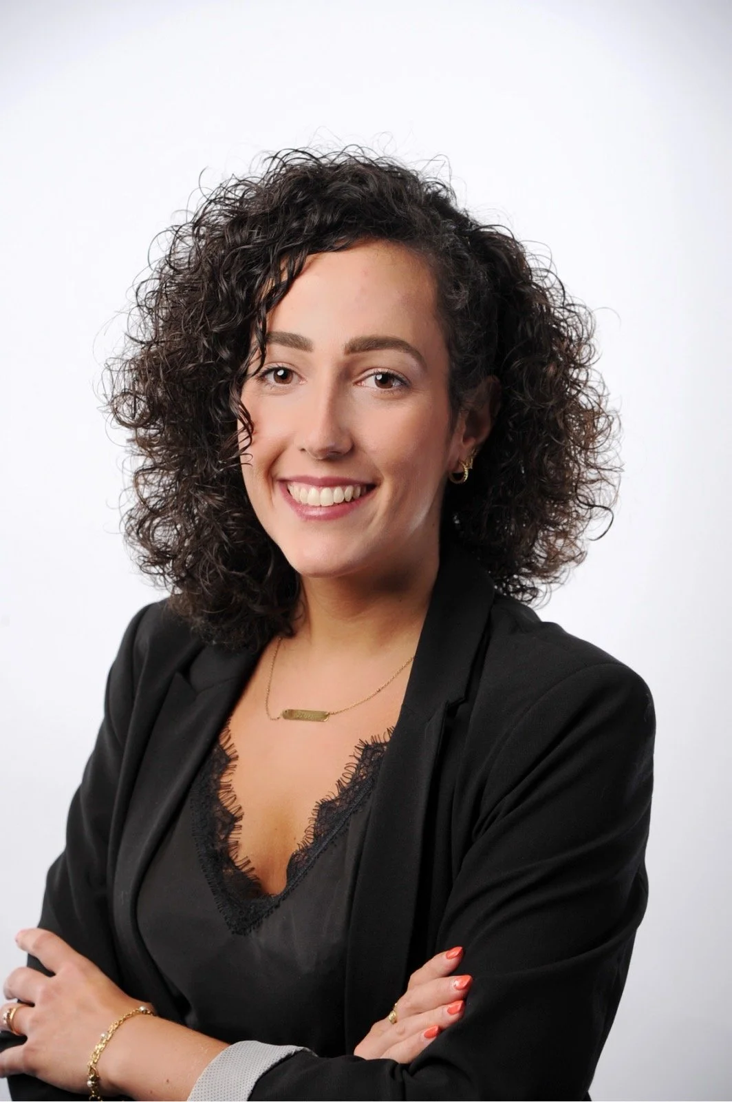 Professional woman with curly dark hair, wearing a black blazer, black lace top, gold jewelry, smiling with crossed arms in front of a plain white background.