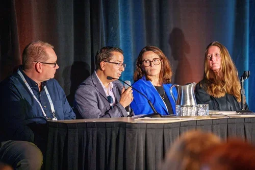 A panel of five diverse individuals sitting at a long table on a stage during a conference or panel discussion. The table has name tags and microphones in front of each person, with an audience visible in the foreground.