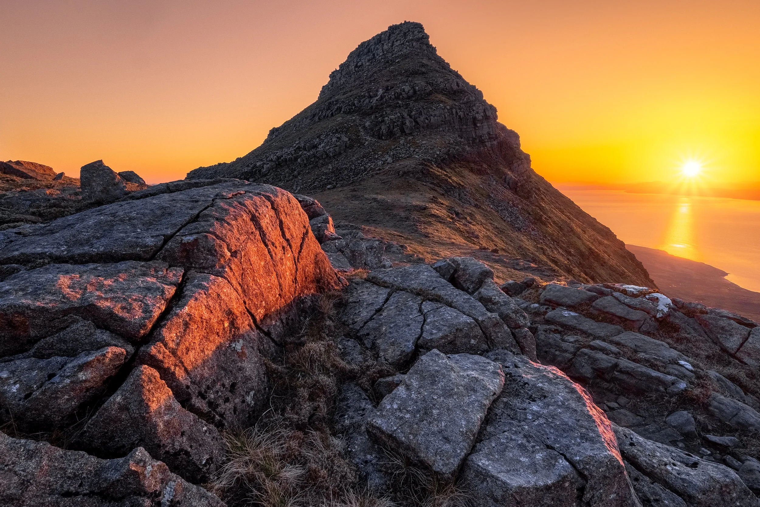 Woman in a light green jacket, gray beanie, and black gloves holding a camera in a rocky outdoor landscape with mountains in the background.