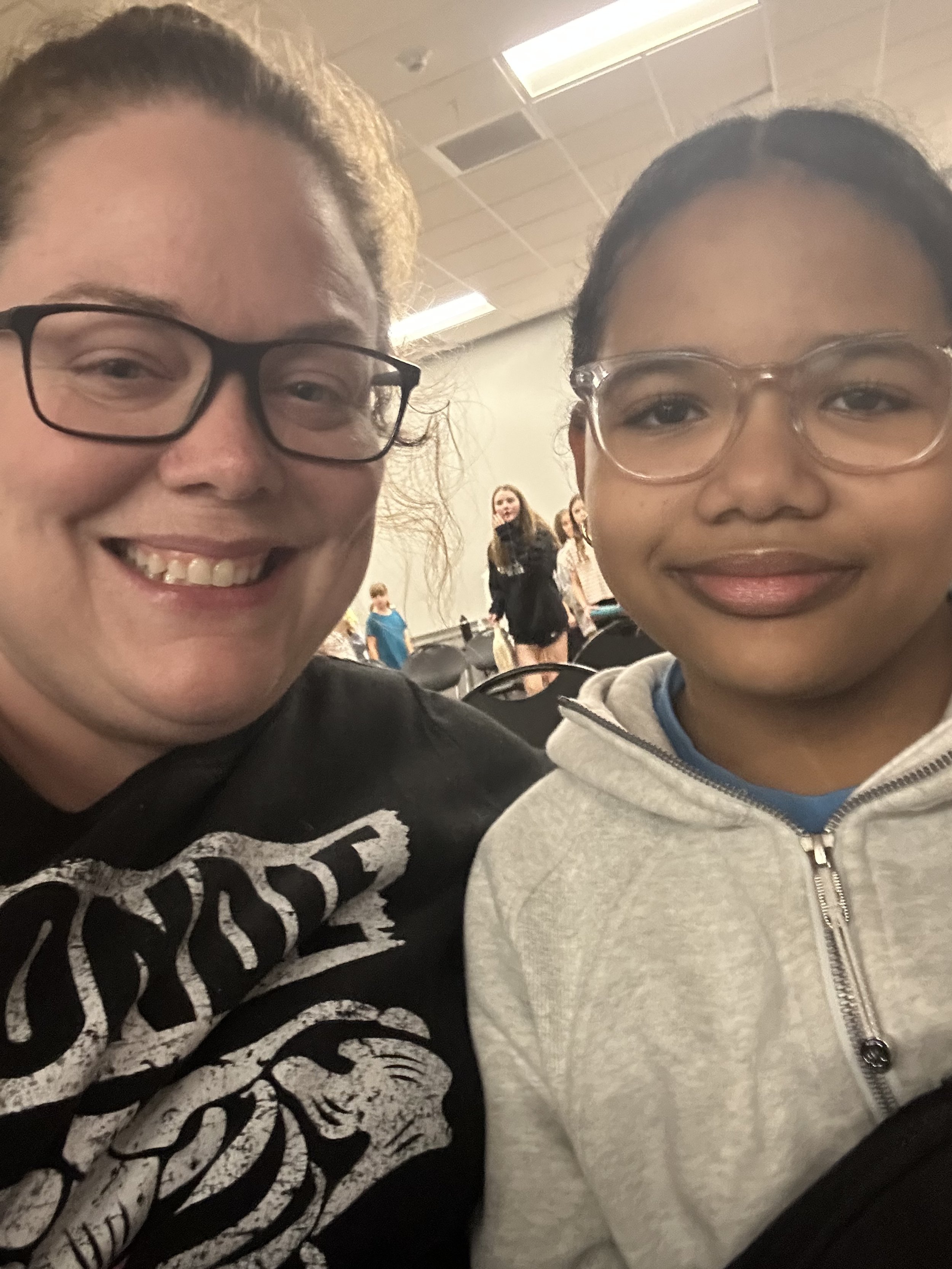 Two people smiling indoors, wearing glasses, with a blurred background of a room with other people.