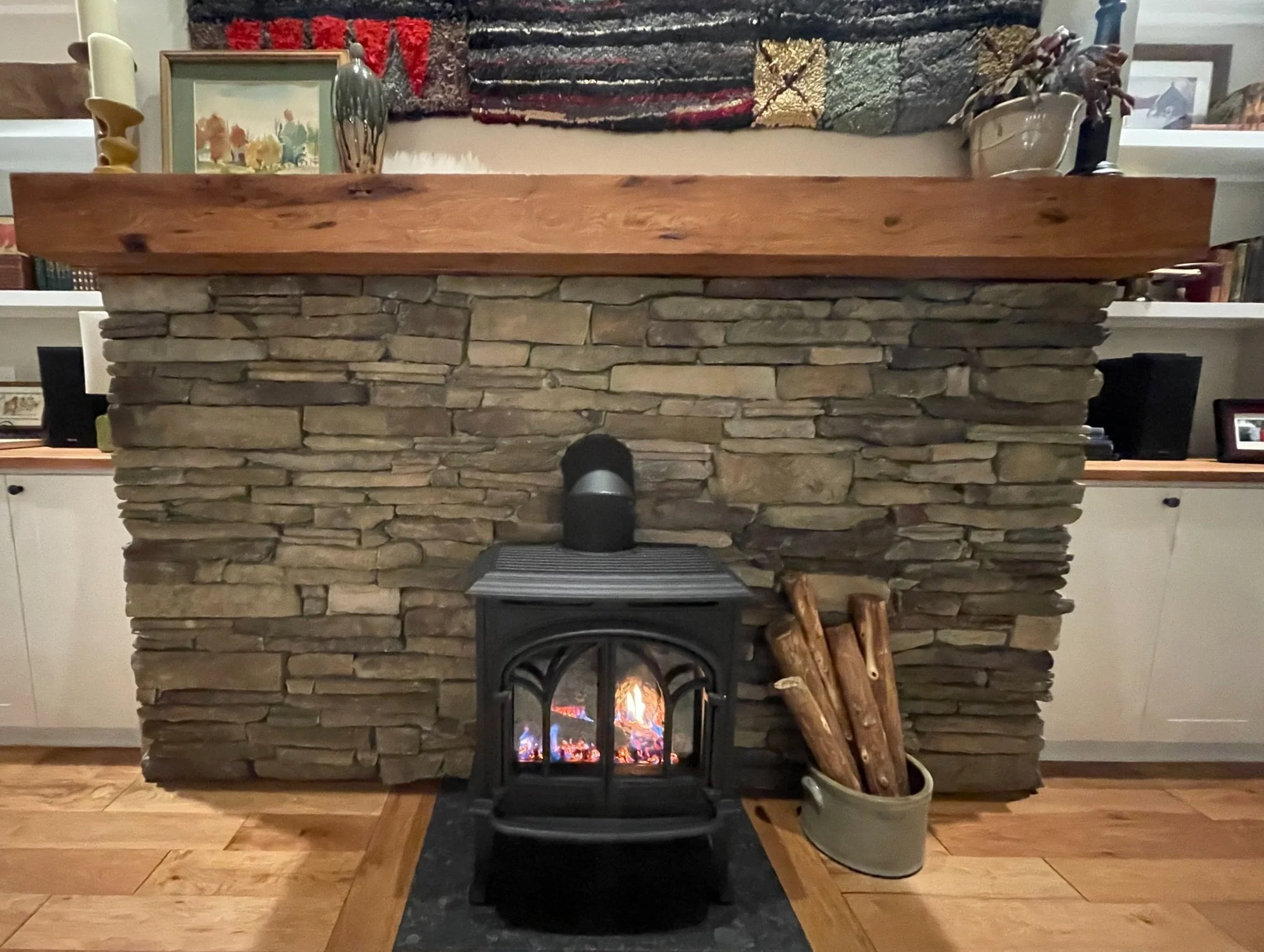 A cozy stone fireplace with a wooden mantel, a small stove with a fire inside, next to a container of firewood, surrounded by a wooden floor and framed artwork and decorations on the wall.